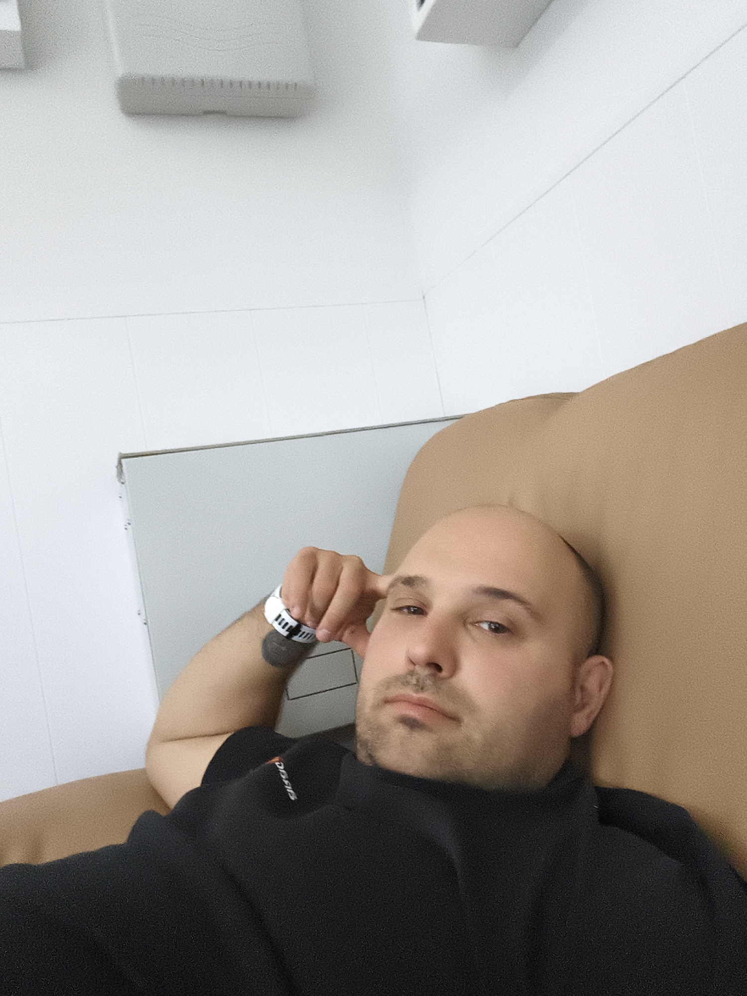 A bald man with a goatee and mustache lies on a tan recliner, looking towards the camera. He wears a black shirt. On his left wrist, a white hospital band is visible. Behind him, a gray metal cabinet and a white tiled wall are present. The room appears to be in a medical setting, possibly a clinic or hospital.
