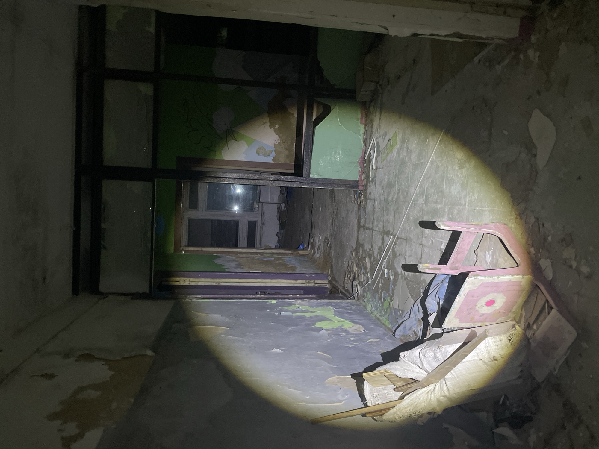 In an abandoned building, possibly a former kindergarten or children's facility, in the village of Yekaterinovka, Russia, a flashlight illuminates a debris-strewn hallway. The walls are peeling, revealing layers of paint in various states of decay, with a prominent green section on the right side bearing faint, abstract drawings. A doorway ahead leads to another room with windows. In the foreground, a pink wooden child's chair, overturned and damaged, rests amidst scattered debris, including sacks and wooden planks. The floor is cracked and littered with rubble. The scene suggests neglect and the passage of time, typical of abandoned structures.