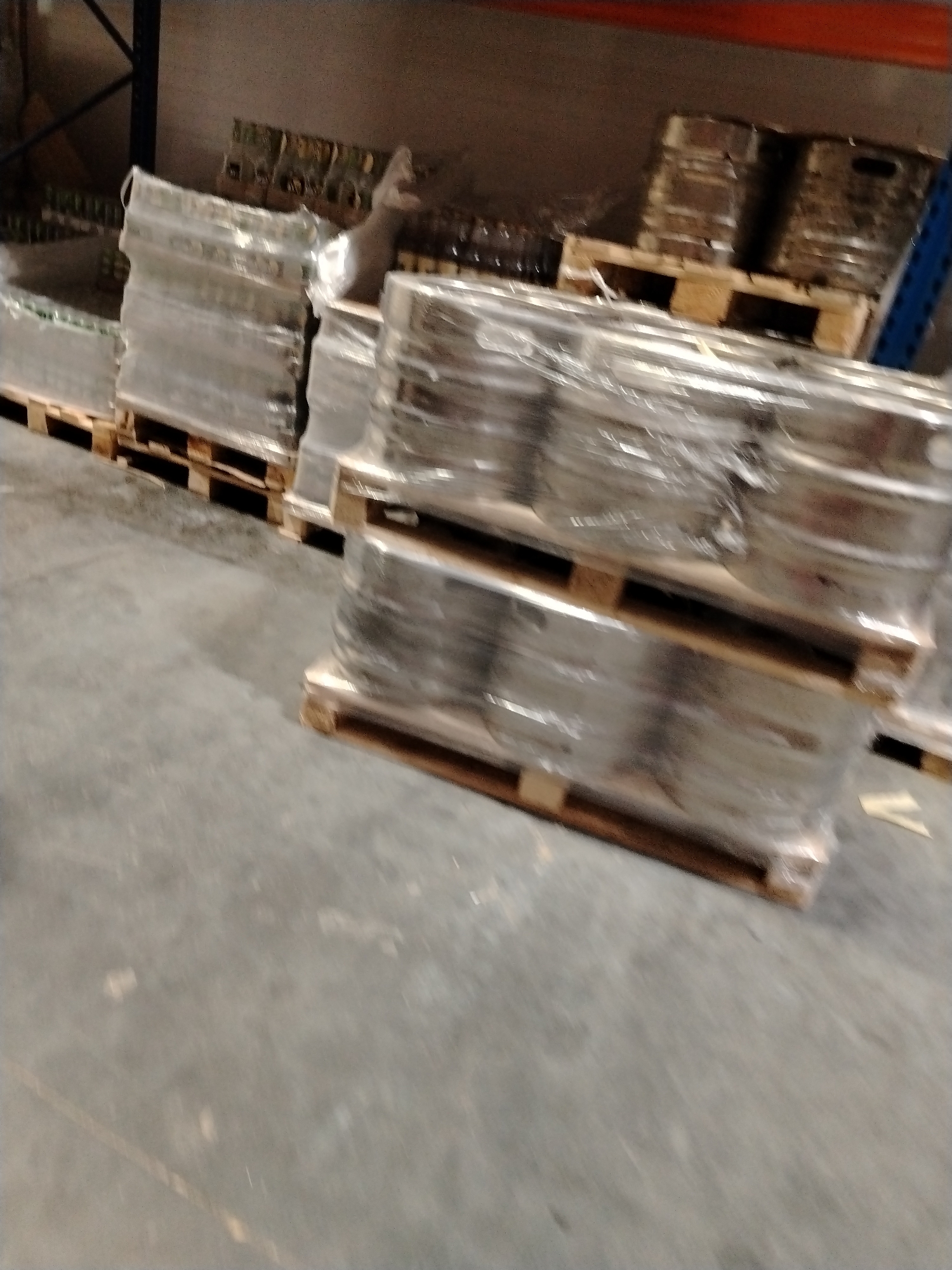 The image captures an indoor scene within what appears to be a warehouse or storage facility, possibly in Rusanovo, Russia. The primary subjects are numerous metallic containers, likely kegs or barrels, stacked on wooden pallets across a concrete floor. Many of these stacks are wrapped in clear plastic film, suggesting preparation for storage or shipment.
In the foreground and midground, several pallets are laden with these shrink-wrapped metallic containers. A prominent stack of at least two layers of containers is visible on the right, with more stacks extending into the background. To the left, another pallet with similar containers is partially visible. The containers appear to be made of a reflective metal, possibly stainless steel.
The background reveals more storage, including industrial shelving units, one of which features a distinctive blue vertical support on the far right. Additional pallets holding what seem to be other packaged items or boxes are also visible on these shelves. The floor is made of concrete, showing some signs of wear and minor debris.
No individuals are present in the image, and no specific activity is taking place; the scene primarily depicts goods in storage. The lighting suggests artificial illumination typical of an industrial setting. The image is somewhat blurry, particularly in the background, obscuring fine details. There is no visible text or identifiable brand information on any of the objects. The overall atmosphere is that of an organized, industrial storage environment.
