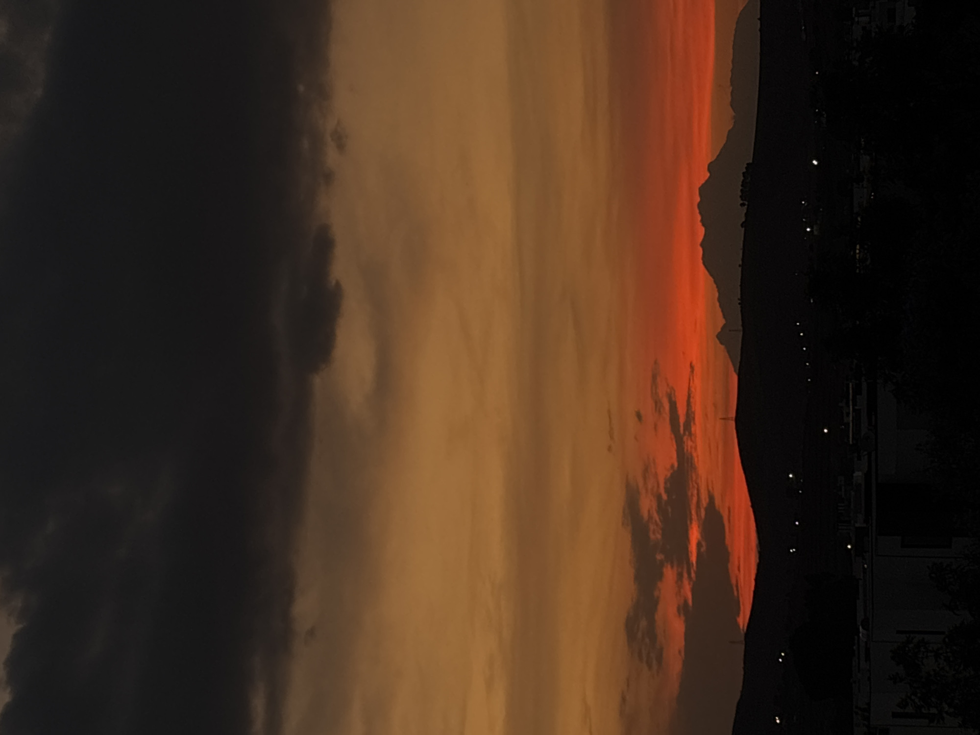 The image depicts a sunset in Santa Clara Ocoyucan, Mexico. The sky transitions from dark clouds on the left to orange and red hues on the right, indicating the setting sun. A silhouetted mountain range is visible against the bright sky. On the right, buildings and trees are visible, lit by scattered lights, suggesting an urban area.