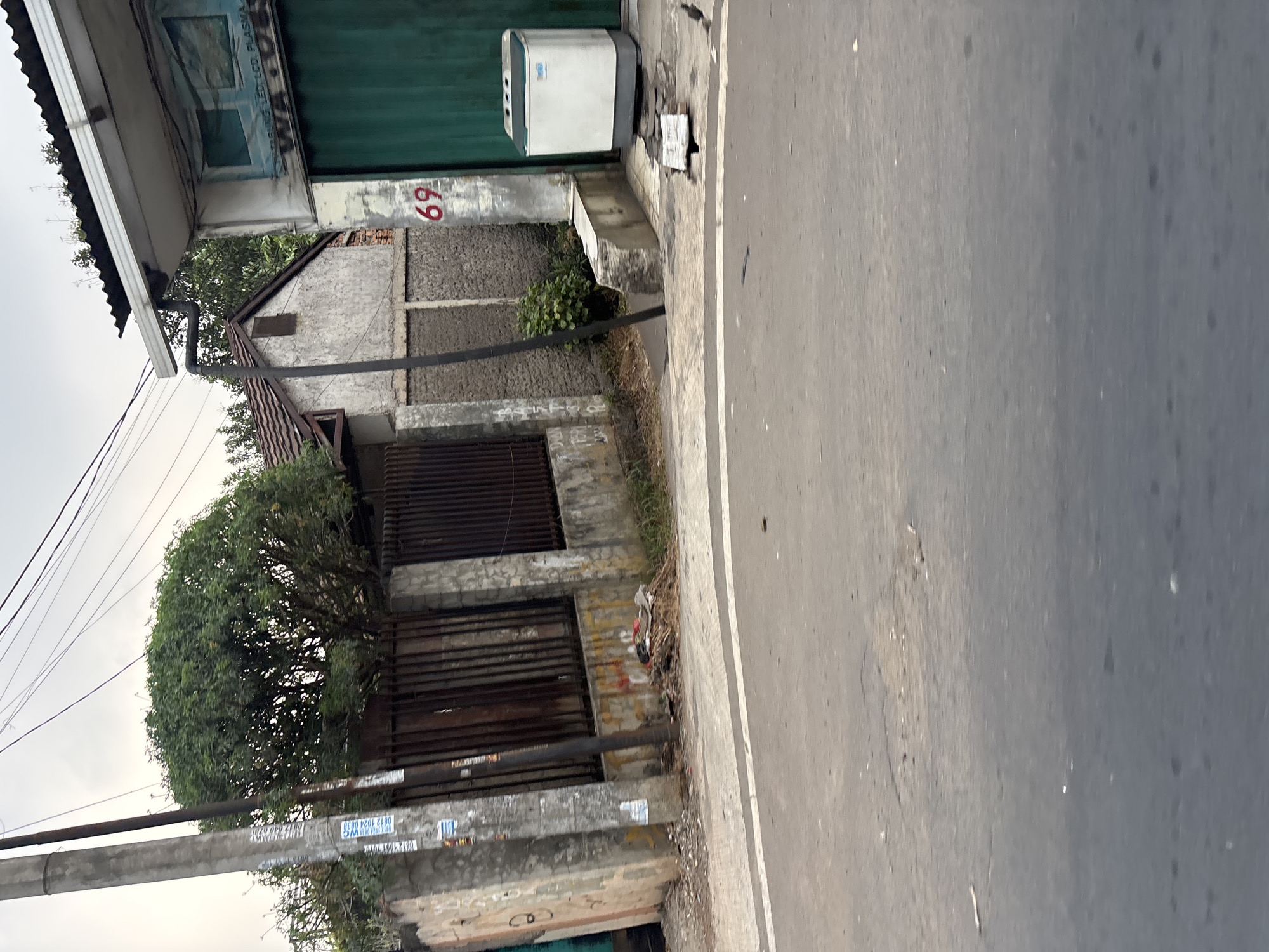 The image captures a scene on a street in the city of Bekasi, Indonesia. In the foreground, a gray asphalt road with a white line marking runs across the bottom of the frame. The road appears somewhat worn with minor cracks and debris.
To the left of the road, a concrete wall with a metal fence runs along the property line. A utility pole stands prominently next to the wall, adorned with several stickers and what appears to be signage. Behind the fence, a building with a textured, light-colored facade and a dark tiled roof is visible, partially obscured by a large, leafy tree.
To the right of the fence and building, a modern structure with a corrugated metal roof and a large, green rolling door is present. A white and gray washing machine sits on the sidewalk in front of this structure. Adjacent to the green door, on a concrete pillar, the number "69" is painted in red. A light-colored concrete bench is also visible in front of this pillar. The sky above is overcast, suggesting cloudy weather. The overall environment appears to be a mixed residential and commercial area.