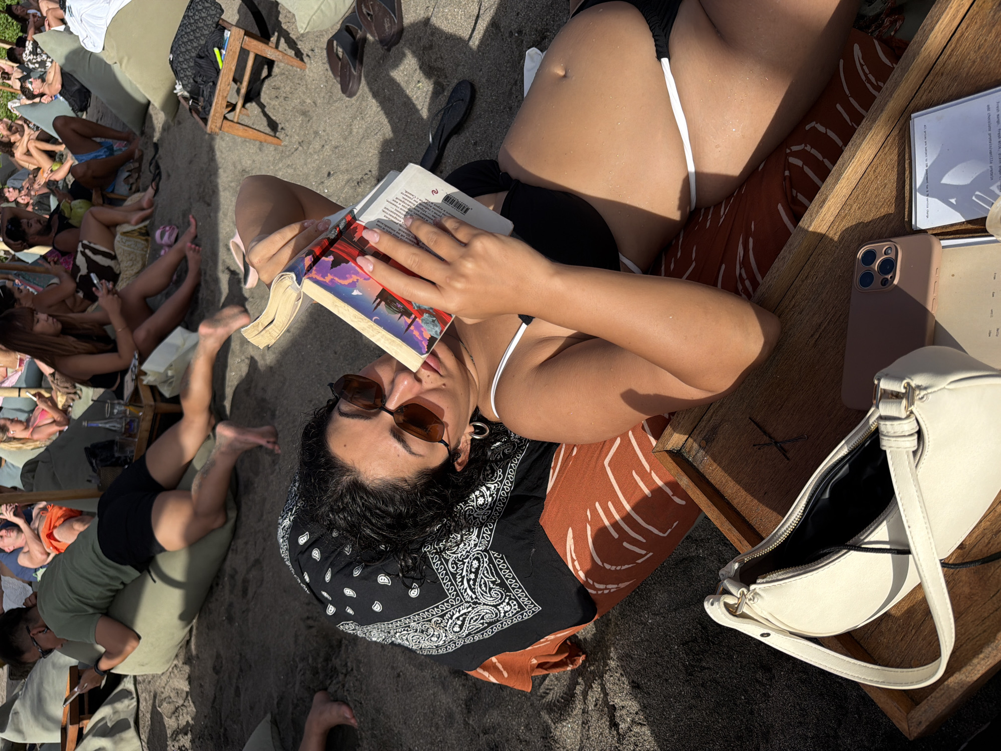 In North Kuta, Indonesia, on a sandy beach, a woman is lying on a cushioned lounge chair, reading a book. She is wearing a black bikini top and sunglasses, and her hair is dark and curly. A white handbag and a smartphone are on a wooden tray next to her. The background shows other beachgoers sitting on beanbag chairs, some also reading or looking at their phones. The sun is shining, casting shadows on the sand.
