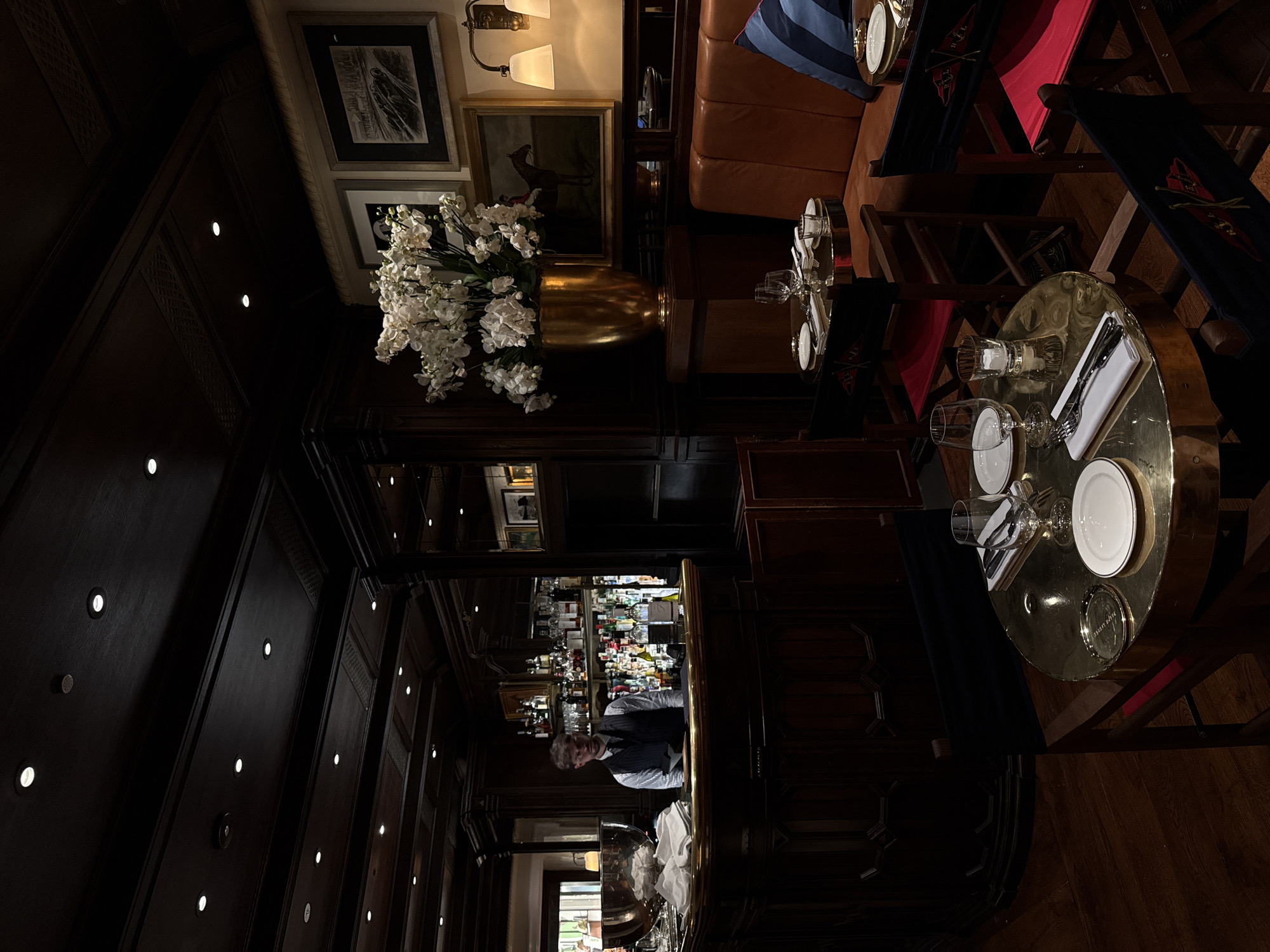 The image depicts an interior scene, likely a high-end bar or restaurant, possibly in Milan, Italy, based on the aesthetic. The room has dark wood paneling with recessed lighting on the walls and ceiling. A large floral arrangement sits atop a dark wooden structure, possibly a room divider or part of the bar. Several framed pictures adorn the walls. The bar area is staffed by a person in a white shirt and dark vest, visible behind a well-stocked bar. The seating area features leather banquettes, red dining chairs, and small round tables set with plates, glasses, silverware, and napkins. The floor is made of wood. The overall lighting is dim, creating an intimate atmosphere.