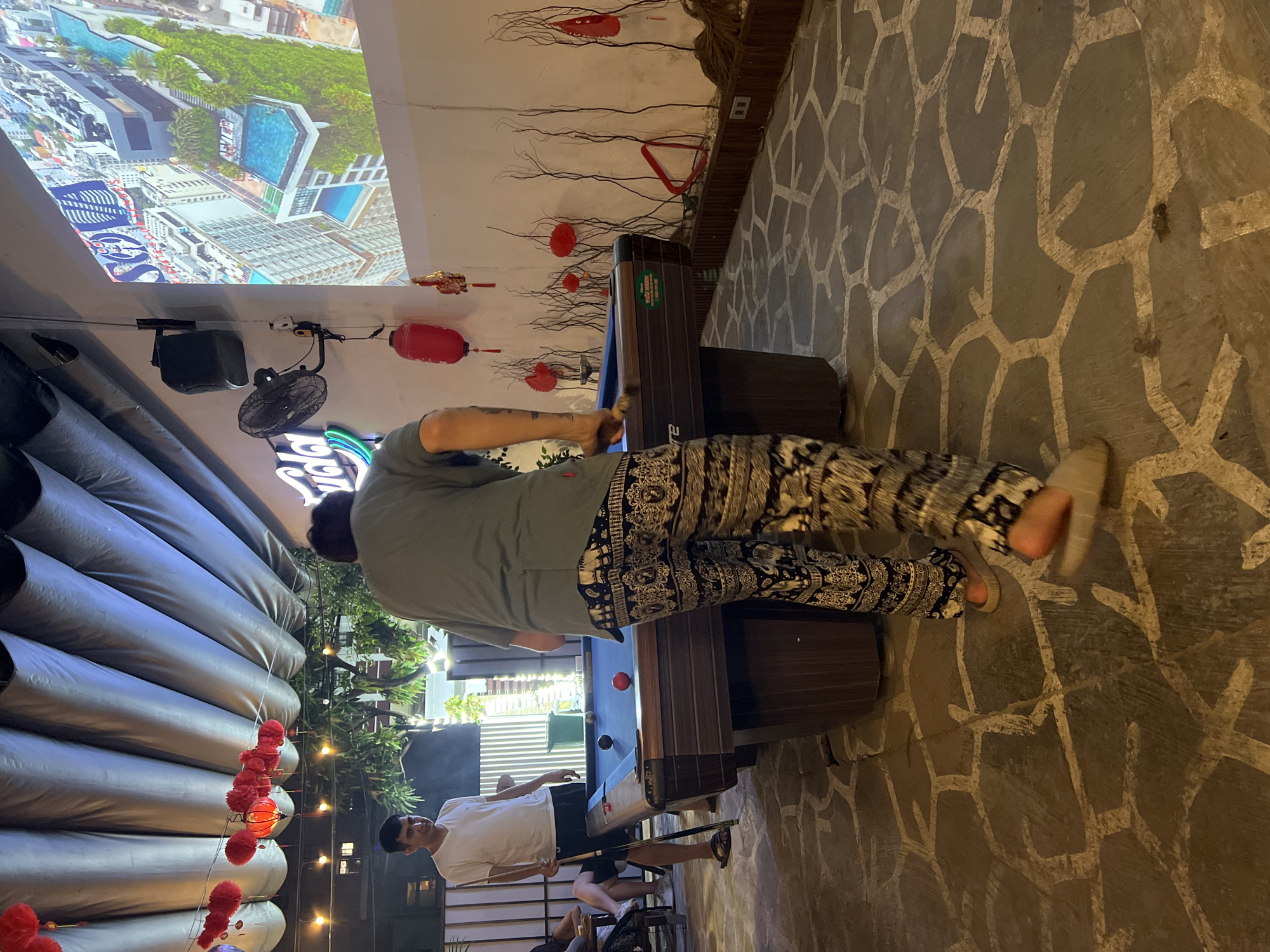 The image shows two men playing pool in what appears to be an outdoor bar or restaurant in Đà Nẵng, Vietnam. One man, wearing a gray shirt and patterned pants, is bent over the pool table, lining up a shot. The other man, wearing a white t-shirt and black shorts, is standing near the end of the table. A projection screen displays an aerial view of a cityscape, possibly featuring hotels or resorts with swimming pools. Red lanterns are hanging as decorations. The flooring is a stone-like pattern. A series of horizontal, silver tubes or cylinders is visible along one side of the space.