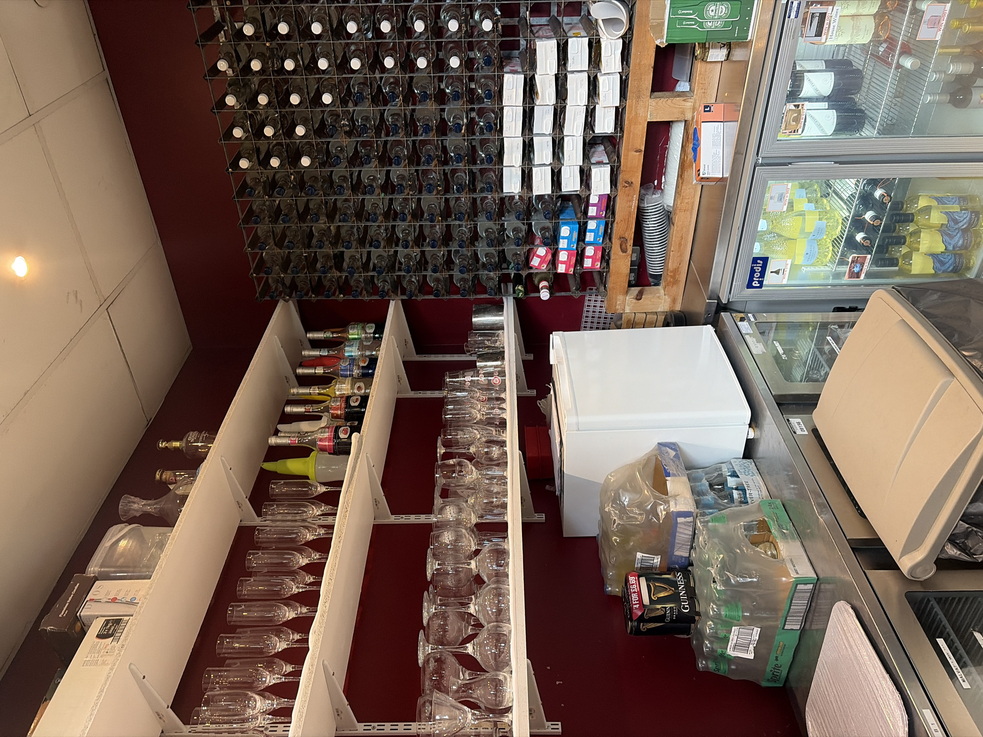 The image displays an interior view of a commercial service area, consistent with a bar or restaurant back-of-house, located in London, United Kingdom. The space features dark red walls and a light-colored ceiling with a single recessed light fixture.
Three white, wall-mounted shelves are positioned along the left wall. The top shelf contains a black box, a clear plastic container, a glass decanter, and two bottles. The middle shelf holds multiple rows of clean, empty champagne flutes, a yellow/green plastic pourer, and several syrup bottles, including one labeled "Aperol." The bottom shelf displays various clean, empty wine glasses and tumblers, with a silver metal container on its right end.
A stainless steel countertop occupies the lower foreground. On the countertop, a pink ribbed mat is visible on the left. To the right, several beverage multi-packs are stacked, including two 4-packs of Guinness stout cans priced "4 FOR £6.99" and a 6-pack of Sprite Zero Lemon Lime bottles. Beneath the countertop on the right, a light beige plastic waste bin is present. Two double-door commercial display refrigerators from the "Prodis" brand are integrated below the countertop, stocked with bottles of white wine, red wine, and other beverages, some labeled "WIN W 001 Home Wines."
The right side of the scene is dominated by a large, floor-to-ceiling metal grid wine rack, filled with numerous clear glass bottles, most with white caps. Its lower sections contain various smaller, colored boxes and white boxed items. Beneath this rack, a structure built from wooden pallets functions as a storage platform, holding rolled-up striped disposable cups, a box of blue disposable gloves, and a green box bearing "Heineken" branding. No individuals or active interactions are visible in the scene.