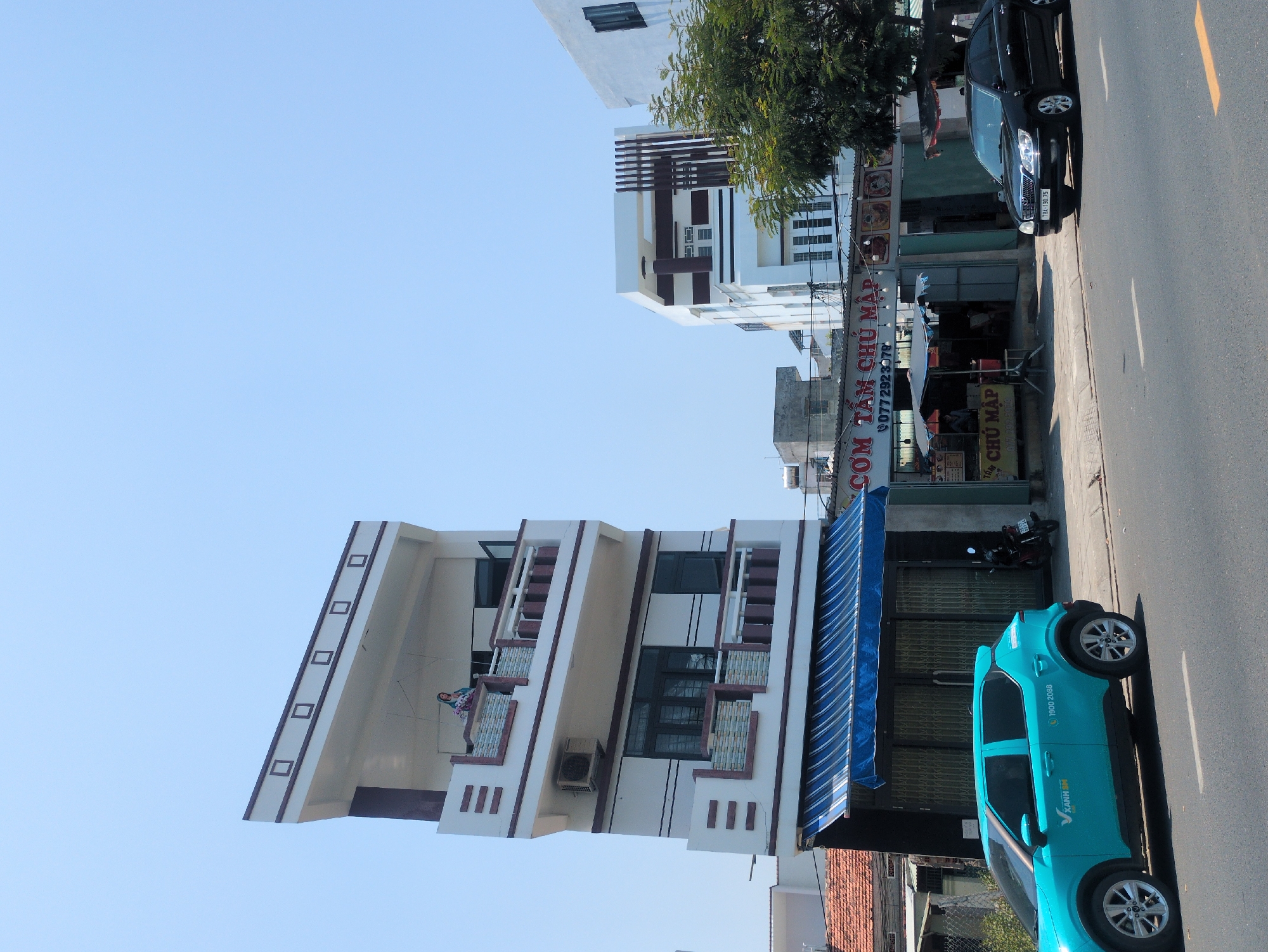 This is a street scene in Tuy Hòa, Vietnam. The image shows a multi-story building with a unique architectural design featuring diagonal elements on its facade. A woman is standing on a balcony of this building. Next to it is a business called "Cơm Tấm Chủ Mập" based on the signs. The building fronting the restaurant has a blue and white striped awning. Parked on the street is a blue car with "XANH SM" markings and "1900 2088" on its side. Further down the street, there's a black car. A motorcycle is also parked near the restaurant. The sky is clear and blue.