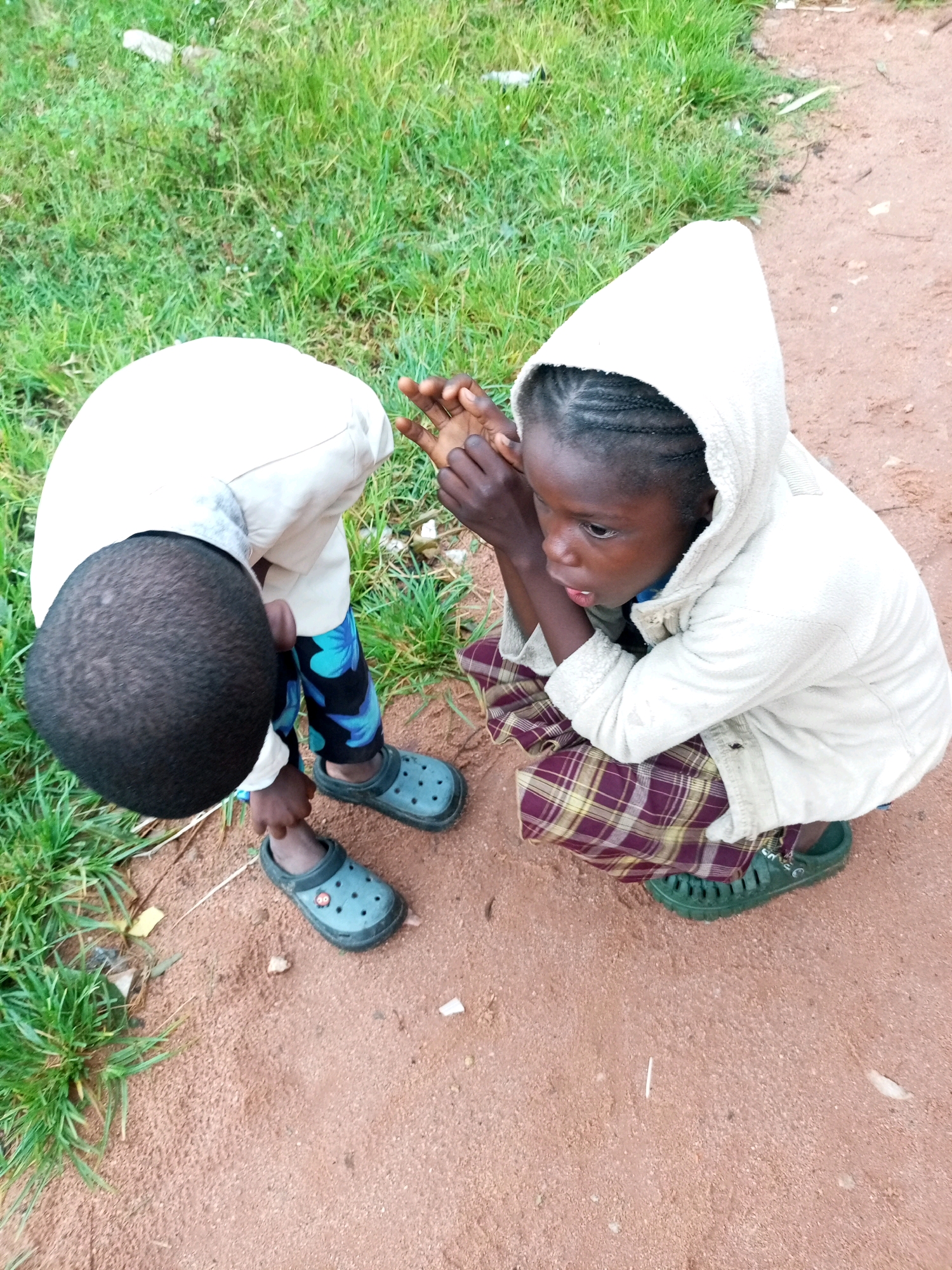 Two young children are crouched on a dirt path, with grass on the left and more dirt path on the right. The child on the left is facing away from the camera, with their head bent down. They are wearing a light-colored hooded jacket and patterned pants with blue and black designs. They have dark, short hair and are wearing dark clogs.
The child on the right is facing the camera and looking down and to the side. They are wearing a light-colored hooded jacket and a plaid skirt with maroon, yellow, and white patterns. Their hair is braided. They are wearing green clogs. Their hands are clasped together in front of them, and they appear to be speaking or listening intently. The environment suggests an outdoor setting, possibly in a rural or undeveloped area. The dirt path is reddish-brown and appears dry. There is some green grass growing along the edge of the path. Scattered on the ground are small pieces of debris, including what looks like white paper or plastic.