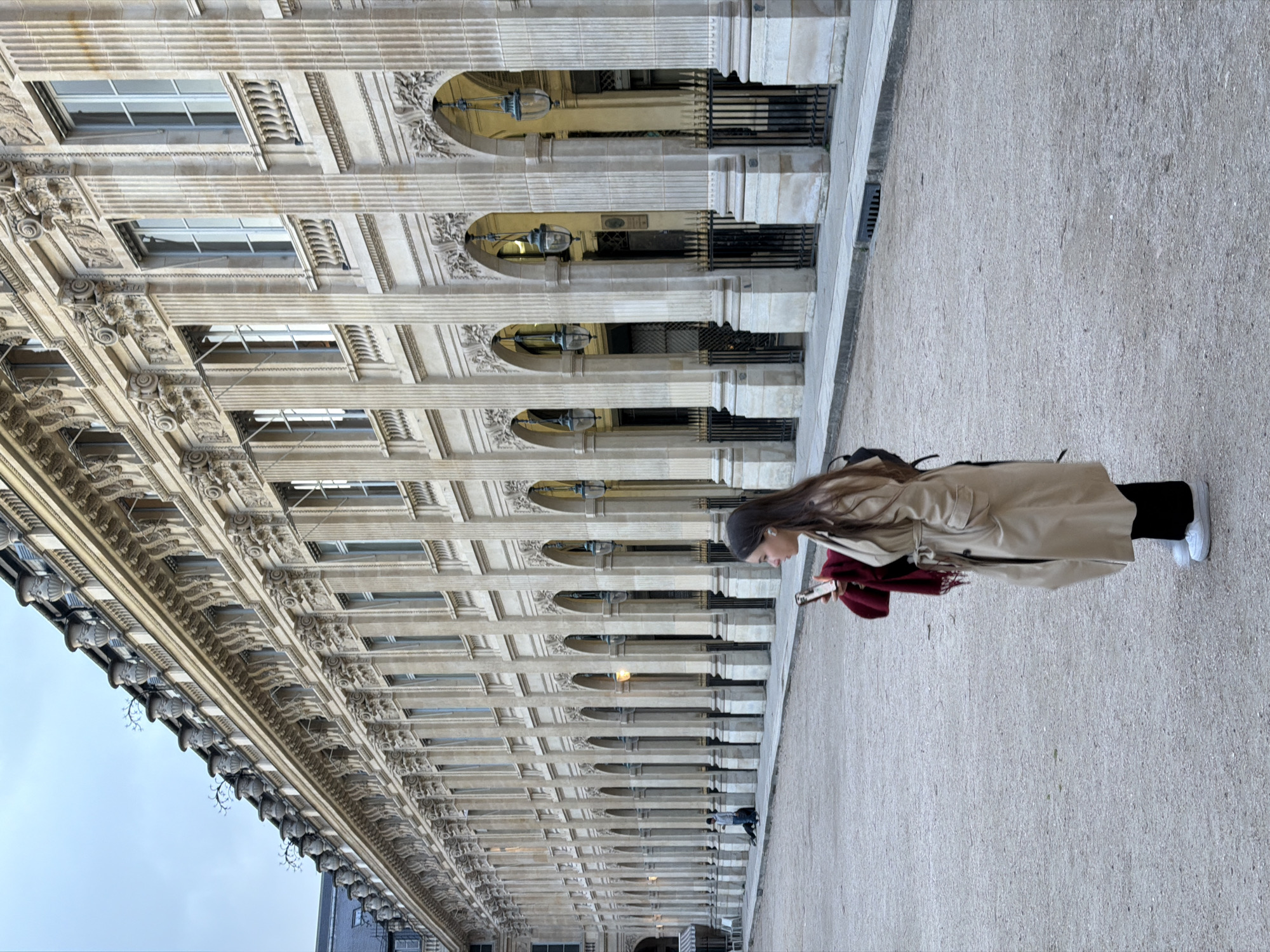 A young woman in a tan trench coat stands in the Palais Royal Garden in Paris, France, looking at a cell phone in her hand. She is facing left. Her dark hair is long and hangs past her shoulders. She is wearing a dark head covering, and her legs are clad in black pants and white sneakers. She holds a red item in her left hand.
The Palais Royal is a formal building in the background of the image, which is oriented on its side. The building has a long arcade with a series of arched openings supported by columns. Windows are visible above the arcade. Elaborate ornamentation adorns the facade of the Palais Royal. The ground is a neutral tone of packed dirt or sand.
