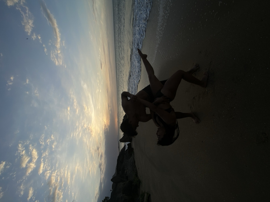 Two silhouetted individuals are depicted on a sandy beach in Juan Griego, Venezuela, at sunset. A male figure, shirtless and wearing dark swim briefs, stands with his body angled towards a female figure. The female, wearing a dark bikini, is bent backward at the waist, supported by or interacting with the male. Her left leg is extended upwards and backward, while her right leg is bent with her foot on the sand. Her head is tilted back, looking towards the male. The foreground is dark sand. To the left, dark rocky formations are visible. To the right, ocean waves gently break on the shore. The sky above features scattered clouds, with warm orange and yellow hues near the horizon, transitioning to light blue overhead. A small bird is visible flying in the upper right portion of the sky.