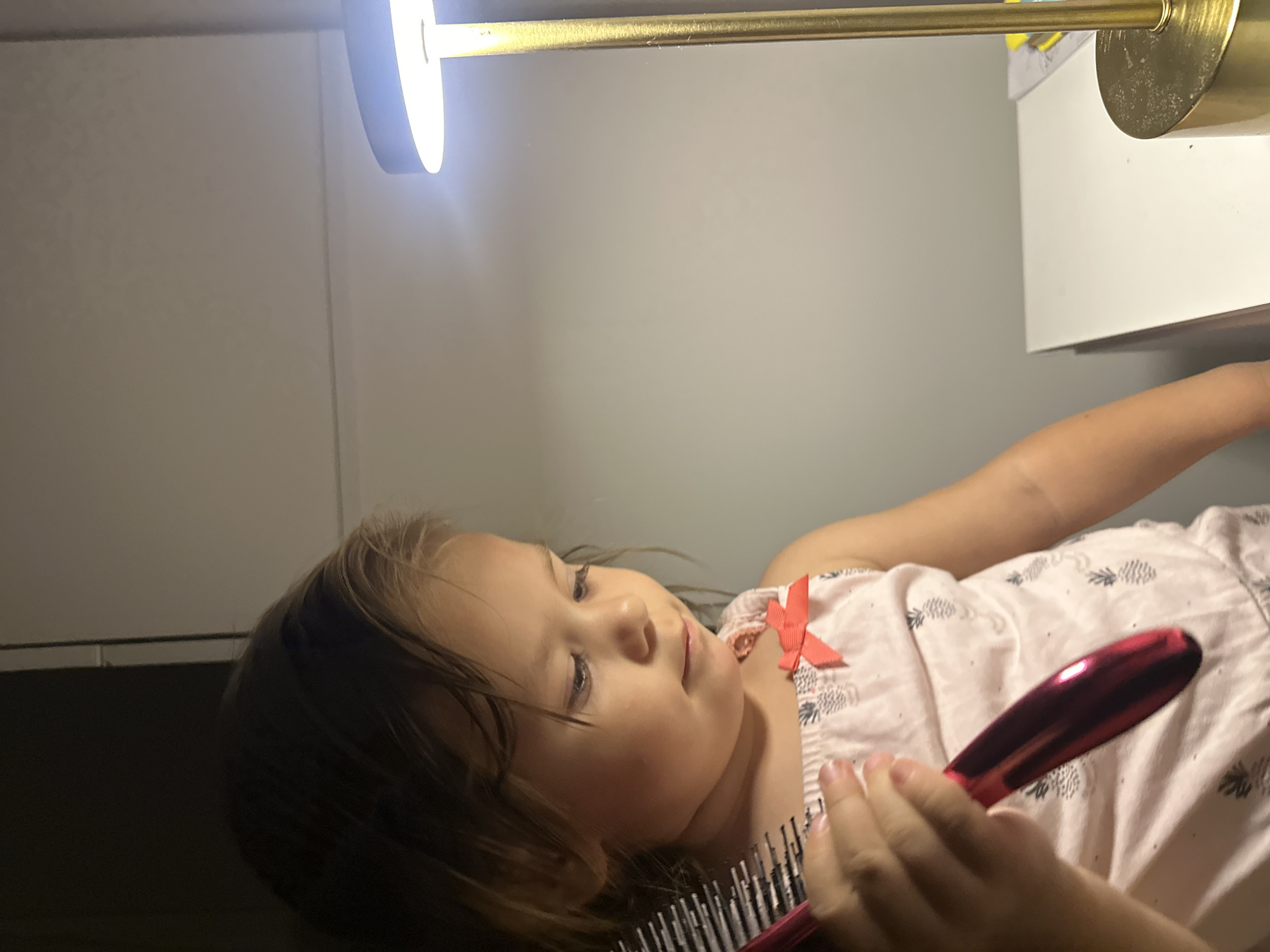 A young girl, approximately three years old, is holding a pink hairbrush in her left hand and looking down at it. She is wearing a light pink sleeveless top with a pineapple pattern and a small red bow on the right strap. The girl has light brown hair and fair skin. She is positioned in front of a light-colored wall, with a portion of a white surface, possibly a desk or shelf, visible to her right. A gold lamp with a bright white light is also present on the right side of the image, casting a glow on the wall. The background to the left of the girl is dark, suggesting a shadowed area. The scene appears to be indoors, possibly in a home or a dressing area. The image does not contain any specific indicators to determine its location within the city of Kharnavy, Russia.