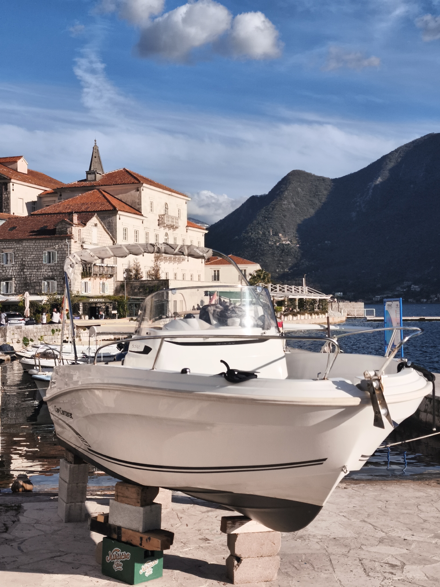 The image shows a white boat on blocks in front of a building and mountain in Kotor, Montenegro. The boat is the main subject, positioned prominently in the foreground and resting on a makeshift support structure of bricks, wooden blocks, and a green plastic crate. The building, with its red-tiled roof and light-colored stone facade, suggests an old European architectural style. The mountain in the background has a dark, shadowy texture. The sky is blue with scattered white clouds. Other boats are visible in the water to the left of the main boat. The overall scene appears to be a harbor or marina on a sunny day.