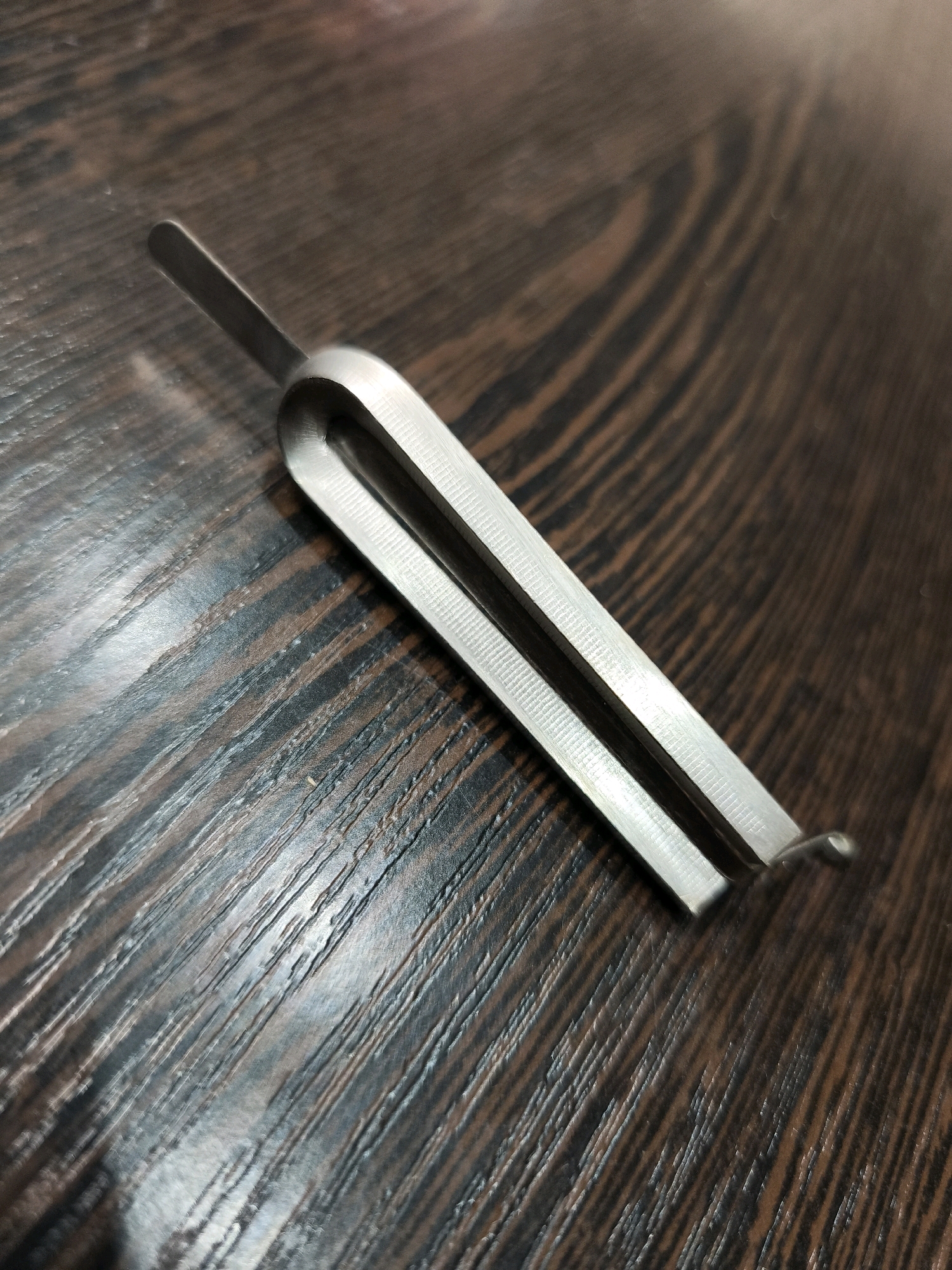 A metal object resembling a tuning fork is positioned diagonally on a dark wooden surface. The object is silver-colored and has a U-shaped head with two parallel prongs. One prong extends further than the other, with a small, rounded tip. The wooden surface exhibits a grain pattern with alternating dark and light brown stripes. The image is an overhead shot, with the object in focus and the background slightly blurred. The scene appears to be an indoor setting, possibly a table or desk, and no specific location in Karachayevsk, Russia, can be identified from the visual information provided.