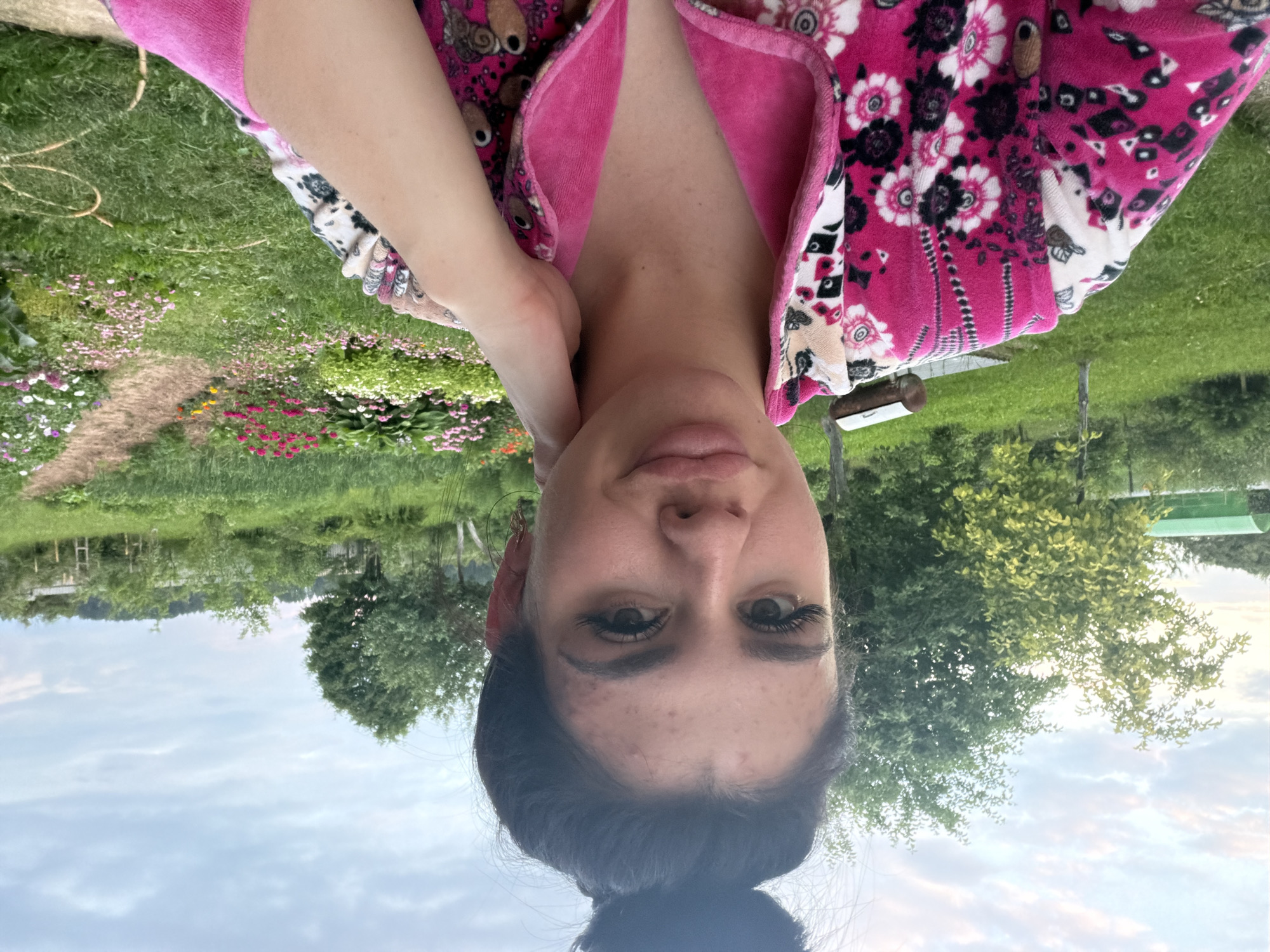 A young woman with dark hair pulled into a bun is taking a selfie outdoors. She is wearing a pink and white floral robe. Her left hand is resting on her neck. The background is a yard with green grass, trees, and a flower garden. A dirt path winds through the garden. There is a green structure and a metal object in the background. The sky is cloudy. The location is the city of Fokino, Russia.