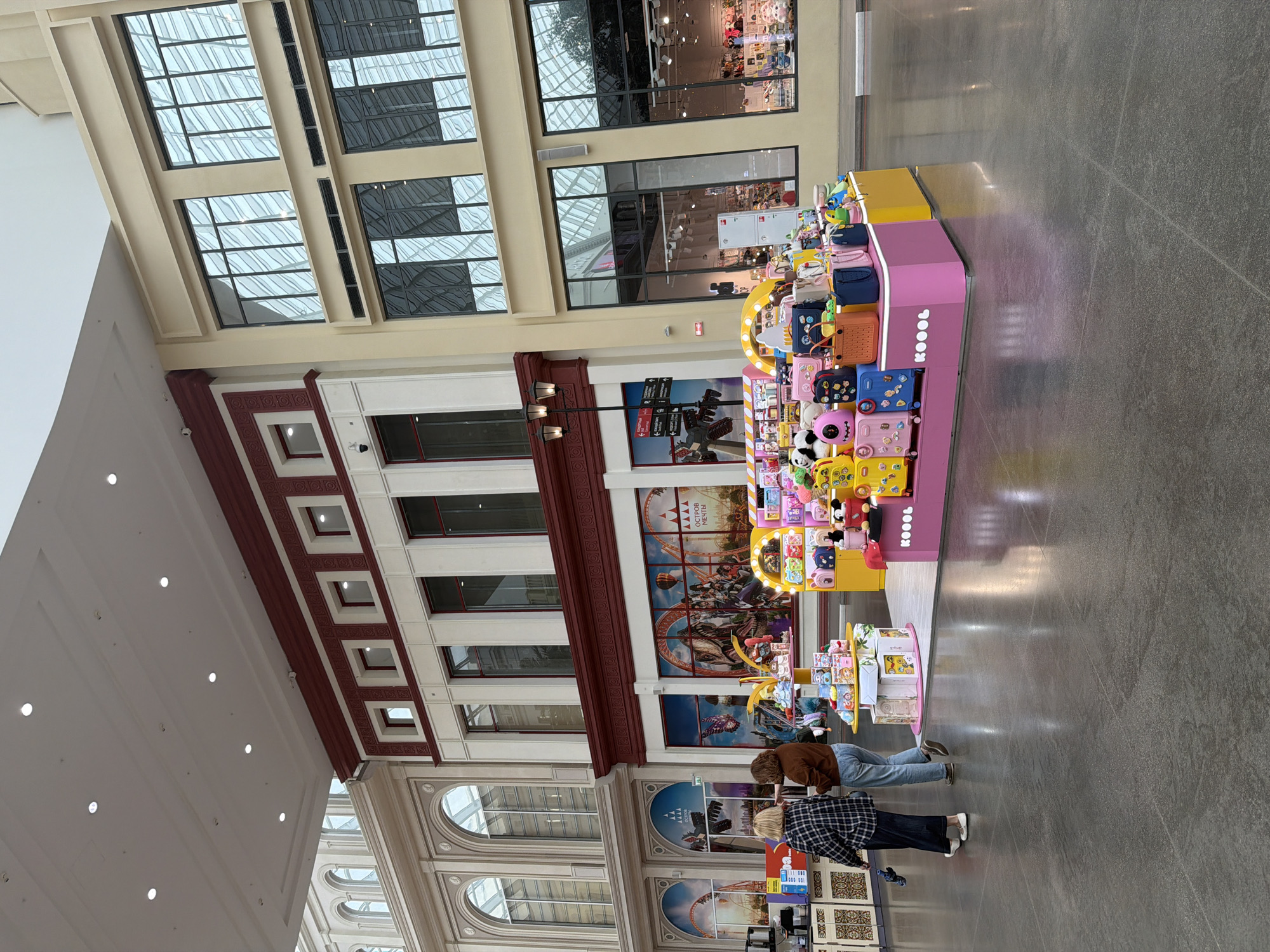 The image captures a wide view of an indoor shopping mall with a faux architectural facade. In the foreground, a brightly colored display of children's luggage and stuffed animals is prominently featured, with the brand name "KOOL" visible on the pink display base. Two women, one in a plaid shirt and dark pants, and the other in a brown jacket and jeans, are walking away from the display towards the left of the frame.
The mall's interior design mimics the exterior of buildings, with cream-colored walls and dark red accents. Large, multi-paned windows are visible on the upper levels of the faux buildings, reflecting the indoor lighting and sky. In the background, the large windows of the shops show merchandise. The floor is polished concrete, reflecting the overhead lighting. The overall impression is a modern, indoor retail environment designed with a theatrical aesthetic.