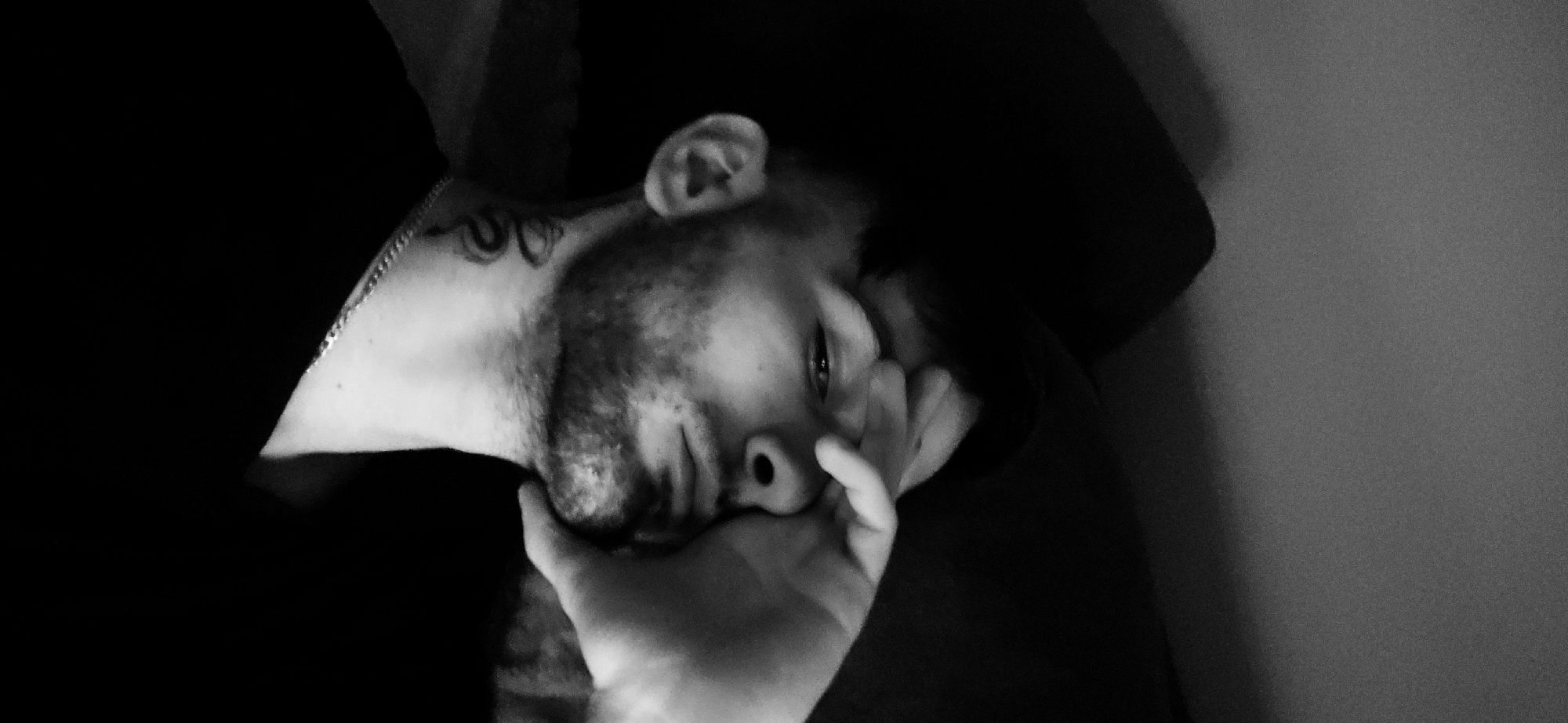 A black and white, close-up photograph depicts a man reclining with his head resting on his hand. The subject has a beard and a tattoo on his neck. He appears to be wearing a necklace or chain. The background is a mix of dark areas and a plain, lighter wall. The location cannot be determined as Plovdiv, Bulgaria, based solely on the visual content provided in the image.