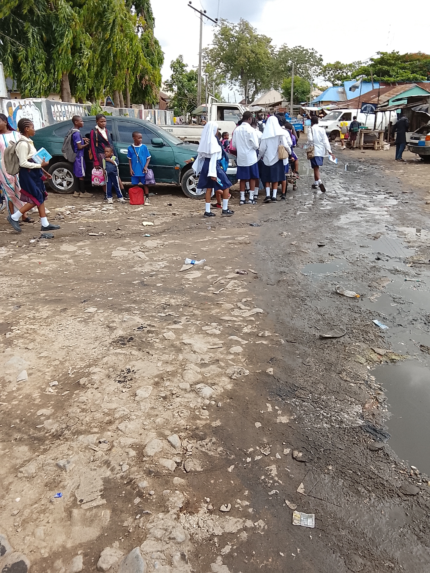 The image depicts a muddy, uneven, unpaved street in Abuja, Nigeria. A group of school children in white and blue uniforms are gathered near a dark green sedan. A few other children and adults are nearby. Debris and trash are scattered on the ground. Across the street, several informal structures with corrugated metal roofs are visible. A couple of other vehicles are parked along the street. The overall environment suggests a low-income or underserved area.