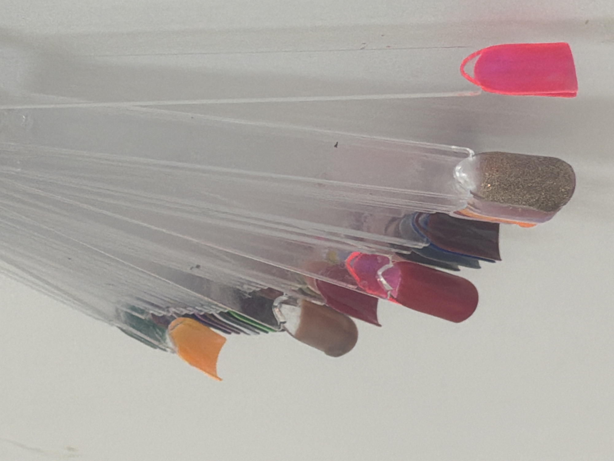 In an indoor setting against a neutral, slightly textured wall, a clear plastic fan of nail polish samples is displayed, hanging vertically. The fan consists of multiple individual plastic strips, each tipped with a polished nail shape showcasing a different color or finish. Starting from the top left, the visible samples include an orange tip, followed by several dark green, purple, and black tips. Moving down the fan, a medium brown tip is visible, then a deep red tip with a subtle shimmer. Further down, a tip with a sparkly gold glitter finish is prominent. To the right of this, a bright neon pink nail sample is attached to a separate plastic strip, hanging lower than the main fan. The lighting is diffuse and appears to be coming from the front, casting soft shadows. The overall impression is one of a nail salon display or a personal collection of nail polish testers. The location is unknown.