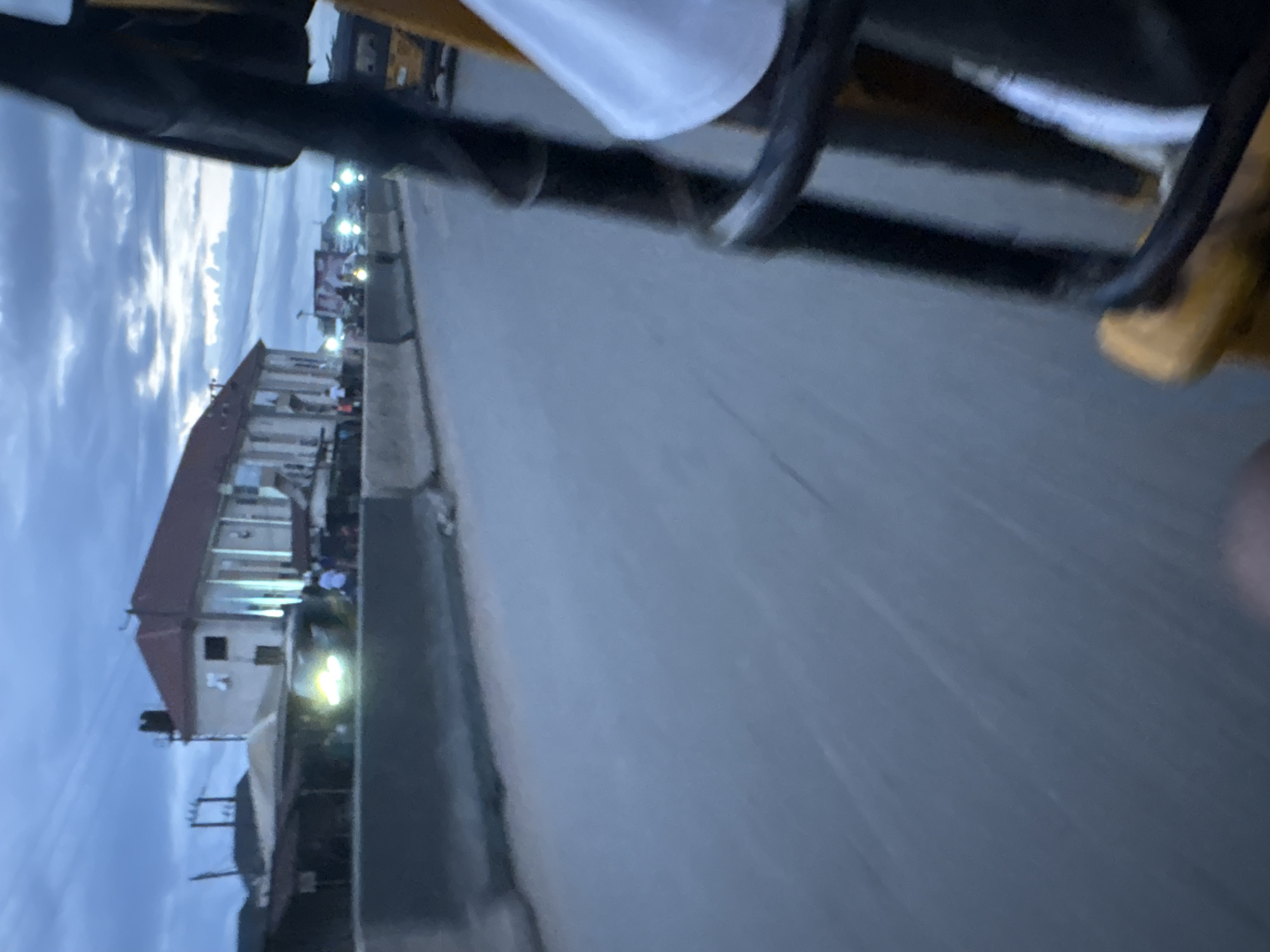 The image shows a view from a vehicle, likely a tricycle, looking towards a road in Onitsha, Nigeria. The sky is overcast and a dim twilight, with scattered clouds. On the left side of the road, there is a multi-story building with a reddish-brown roof. In front of the building, several people are visible, and some vehicles are parked or moving, with their headlights illuminated. The road itself appears to be paved, and there's a concrete barrier separating lanes or the road from a sidewalk/area with structures. The foreground is dominated by the vehicle's interior, with parts of its frame and a person's white shirt visible. The overall impression is of a bustling urban environment at dusk, possibly a market area given the presence of people and various vehicles.