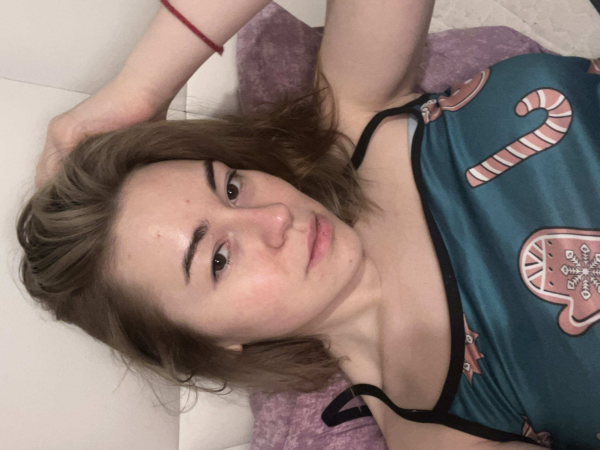 The image displays a young adult female reclining indoors, in a location stated to be Boreti, Montenegro. She possesses light brown, medium-length hair, brown eyes, and a neutral facial expression, looking directly towards the viewer. Her complexion exhibits some redness across her face, notably on the cheeks and forehead, along with a few minor epidermal blemishes. She is clad in a teal-colored camisole with thin black straps, adorned with light pink and white festive designs, specifically a candy cane, a gingerbread man, and a mitten with a snowflake pattern. Her left arm is elevated, with her hand positioned near the top of her head, lightly touching her hair. A red string bracelet is visible on her left wrist. The immediate environment includes a white headboard or wall behind her head, a light purple quilted pillow or blanket to her right side, and a lighter, possibly off-white, quilted bed covering beneath her left shoulder and lower torso. The scene is illuminated by soft, artificial light.