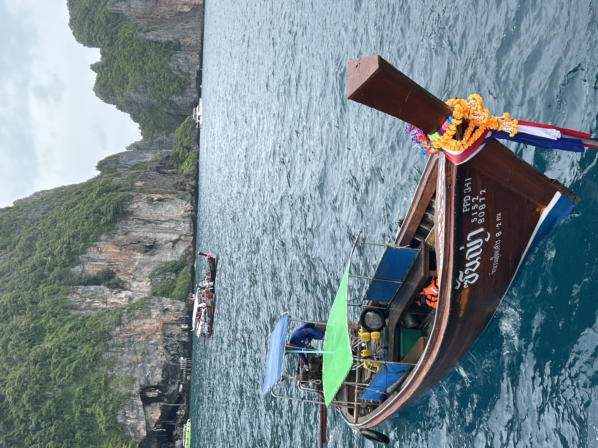 The image captures a longtail boat adorned with floral garlands and Thai flags, bobbing on the turquoise waters of Ao Nang, Thailand. Towering limestone cliffs covered in lush green vegetation rise dramatically from the sea in the background, framing the scene. Several other boats, including a larger passenger boat filled with people, are visible in the distance, suggesting a popular tourist area. The sky is overcast, casting a soft light over the landscape. The longtail boat in the foreground appears to be a tourist vessel, possibly used for island hopping tours. The detailed inscription on the boat, "PPD 341 5152 80872," likely denotes its registration or identifying number. The presence of life vests adds a touch of practicality to the otherwise picturesque setting.