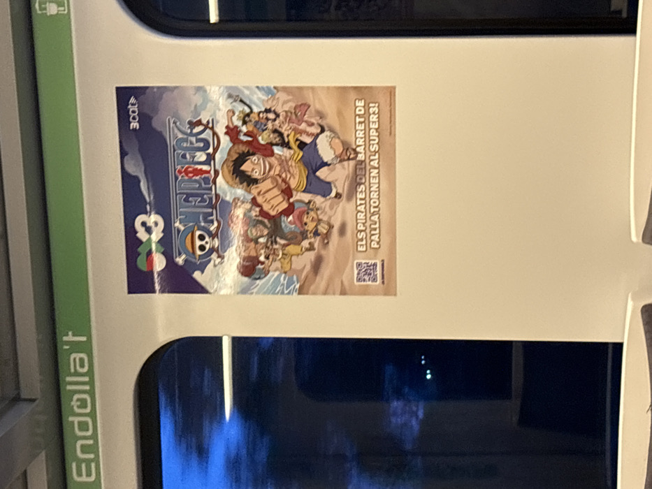 The image displays the interior of a public transport vehicle, likely a train or metro car, situated in Sant Quirze del Vallès, Spain. A large vertical poster, approximately half the width of the wall section, is affixed to a plain white interior panel. The poster features characters from the *One Piece* anime series; Monkey D. Luffy, wearing a straw hat, is central with his right fist extended, surrounded by other members of the Straw Hat Pirates. The *One Piece* logo is displayed at the top center of the poster, flanked by a "3x3" logo on the left and a "3cat" logo on the right. Below the character illustration, Catalan text reads "ELS PIRATES DEL BARRET DE PALLA TORNEN AL SUPERS3!" (The Straw Hat Pirates return to Supers3!), with a QR code in the bottom left corner. Above the poster, a horizontal green strip runs across the top of the interior space, bearing the white text "Endolla't" (Plug in yourself), indicating a charging facility. To the left of the poster, a rounded window displays a dark, blurred exterior view with faint, distant lights, consistent with a vehicle in motion during nighttime or dusk. Portions of grey upholstered seating are visible at the bottom of the frame.