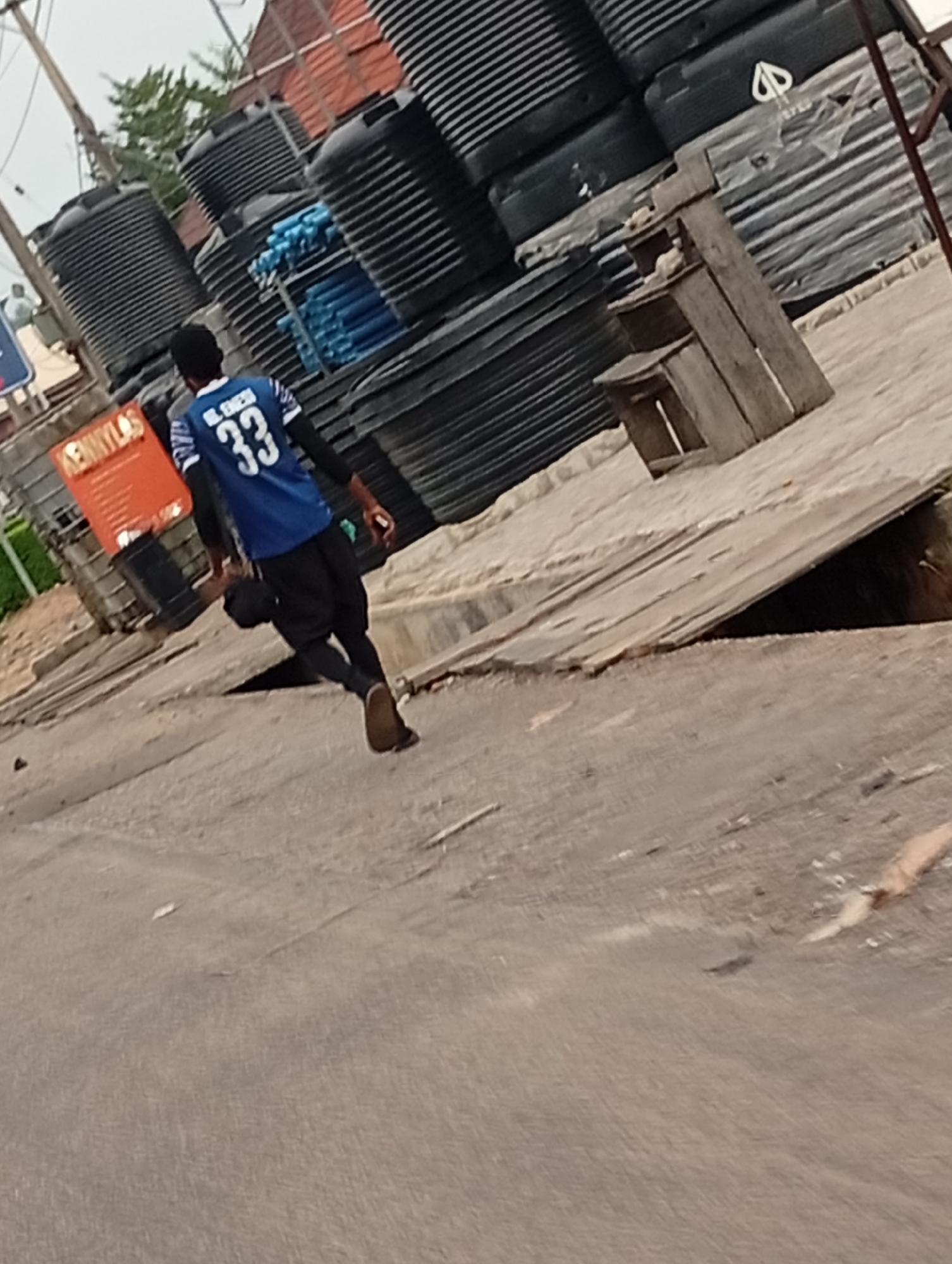 A person wearing a blue jersey with the number 33 is walking on a street in Adankolo, Nigeria. The person is walking on the left side of the street. A stack of large black plastic tanks sits to the person's right side along with some blue piping and a wooden shelf structure. The street is asphalt. An orange sign with white lettering stands behind the person. An open storm drain with a wooden covering is at the edge of the road on the right.