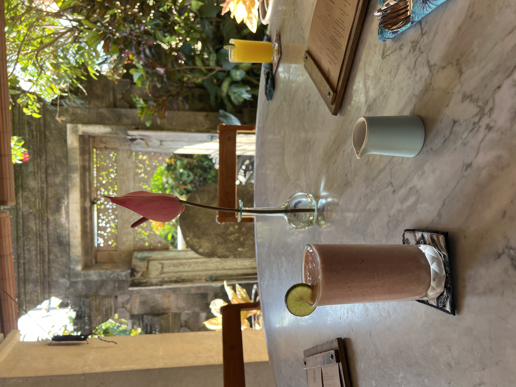 This is a medium shot image taken outdoors at a restaurant in Ubud District, Indonesia. The foreground features a marble-top table with a tall glass of pink smoothie, garnished with a lime slice, placed on a patterned coaster. To the right of the smoothie, a small light blue ceramic pitcher sits next to a menu on a wooden stand. Further to the right, cutlery is neatly arranged on a blue and white patterned cloth. In the mid-ground, a single pink orchid in a clear glass vase stands in the center of the table. Behind the table, an arched opening reveals a lush, green garden with a stone wall and more foliage. Sunlight streams into the scene, creating bright highlights and soft shadows, indicating it is daytime. The overall ambiance suggests a tranquil and inviting outdoor dining experience.