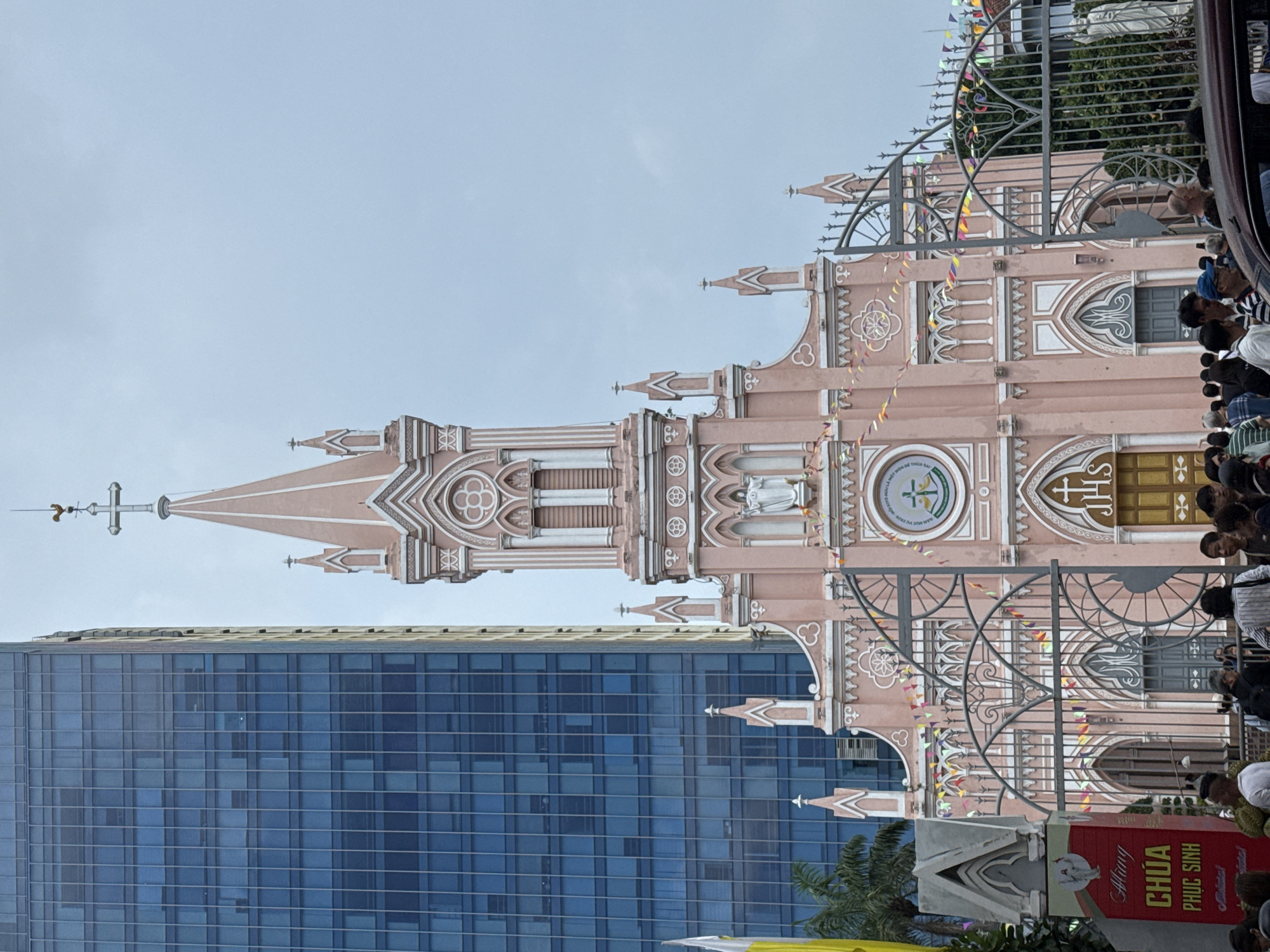 The image depicts the Đà Nẵng Cathedral, a Roman Catholic cathedral in Đà Nẵng, Vietnam. The cathedral, known for its distinctive pink color, features a tall spire topped with a cross. In the background, a modern blue glass-clad high-rise building rises behind the cathedral. A wrought-iron gate partially obscures the view of the cathedral's facade. A crowd of people is visible near the gate, suggesting a gathering or event. A sign in Vietnamese is also visible. The sky is overcast.