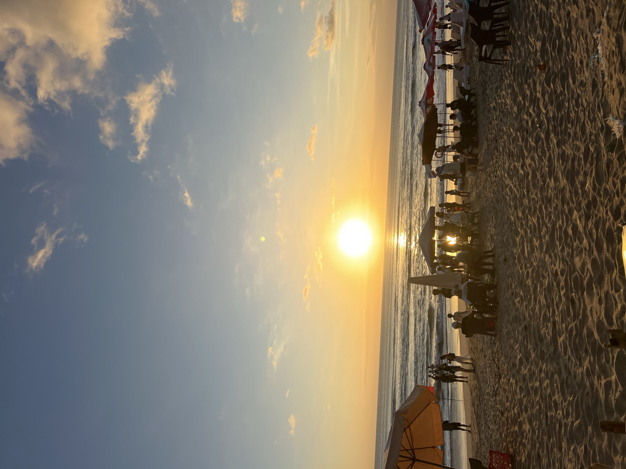 The image depicts a sunset scene at Kuta Beach, Bali, Indonesia. The sun is low on the horizon, casting a warm golden glow across the sky and the ocean. Gentle waves are rolling onto the sandy shore, where numerous people are gathered. Some individuals are standing and walking near the water, while others are seated under beach umbrellas and canopies. Several beach chairs are also visible. The atmosphere suggests a relaxed evening at a popular tourist destination.