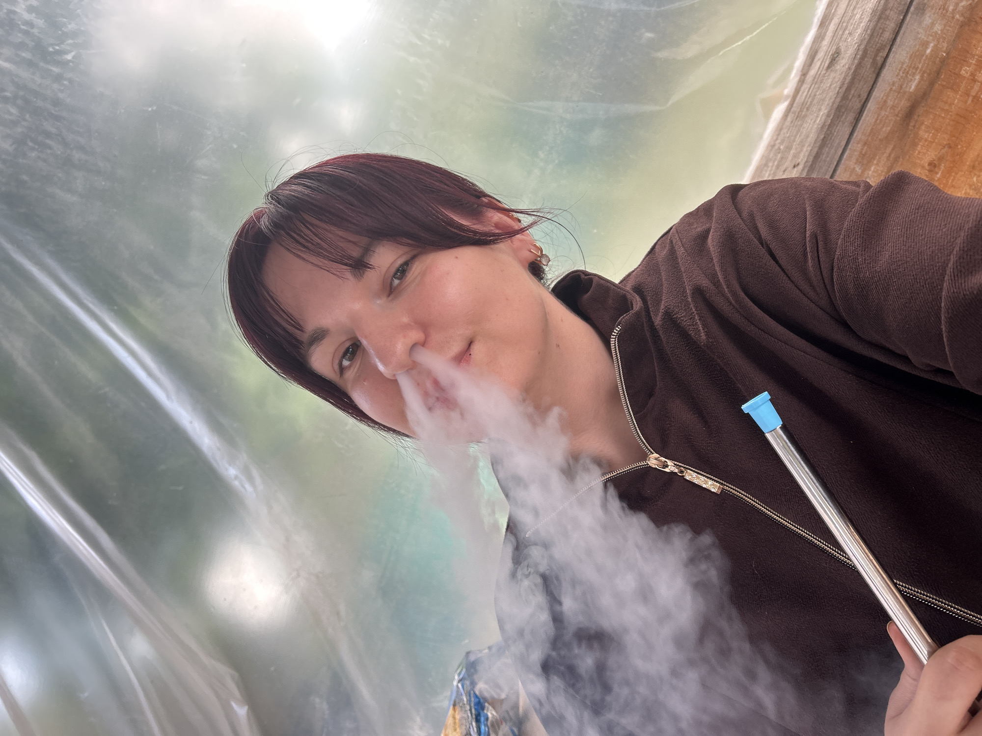 A single female, approximately 20-30 years old, is depicted exhaling a cloud of white vapor from her mouth and nose, holding a metal hookah hose with a light blue mouthpiece in her right hand. She has dark reddish-brown hair with bangs, light skin, and is looking towards the camera with a slight smile. She is wearing a brown textured jacket with a gold zipper. The background consists of clear plastic sheeting, exhibiting wrinkles and reflections, through which a blurry outdoor environment with green foliage and a lighter sky-like area is visible. A wooden surface is present in the lower right corner, and a partially visible object wrapped in foil is on a surface in the upper left background. The scene is located in Фоки, Russia.