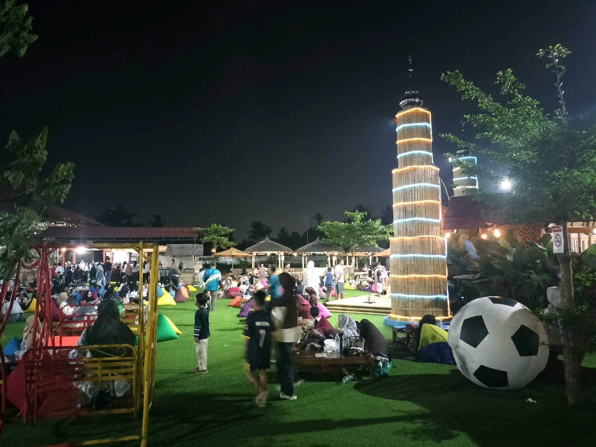 This image depicts a nighttime scene at a recreational area, likely a cafe or park, in Depok, Indonesia. The artificial grass ground is dotted with numerous beanbag chairs in various colors, suggesting a casual and comfortable atmosphere. In the foreground, a red and yellow swing set is present. Several people are seen scattered throughout the area, some sitting on the beanbags, others standing or walking.
Dominating the right side of the image is a large, inflatable soccer ball, a prominent decorative element. Behind it, a tall, cylindrical structure made of bamboo is illuminated with alternating bands of orange and blue lights, giving it a striking visual presence. This structure appears to be a landmark or a key feature of the location. Further in the background, more people are gathered, and there are some open-air structures with thatched roofs, possibly food stalls or seating areas. The overall ambiance is lively, with people enjoying an evening outdoors. The presence of several illuminated towers in the background, similar to the bamboo structure, suggests this might be a themed attraction or a popular gathering spot.