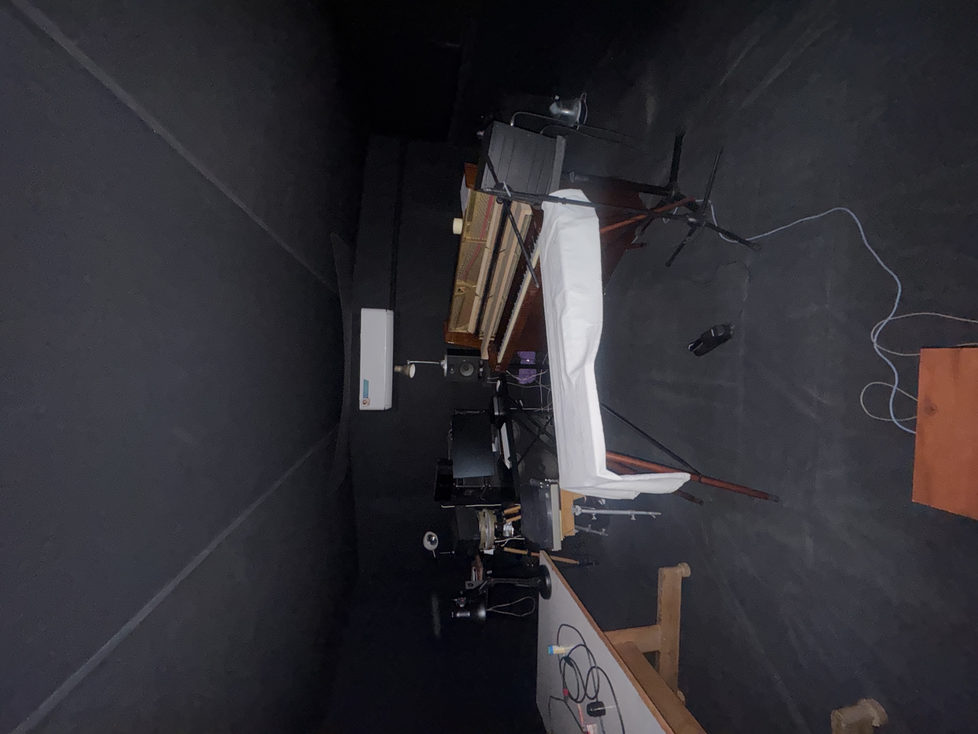 The image depicts a dimly lit interior space, likely a music studio, devoid of any visible people. In the foreground, a light-colored table with cables and electronic equipment is situated on the left. To the right, a piano with its lid open is prominent, covered partially by a white cloth. Behind the piano, a music stand and a chair are visible. The room is characterized by dark walls and ceiling, with some lighting fixtures present. The overall impression is one of a functional yet somewhat cluttered creative space. The location in the city of Gyumri, Armenia, is not discernible from the visual elements within the image.