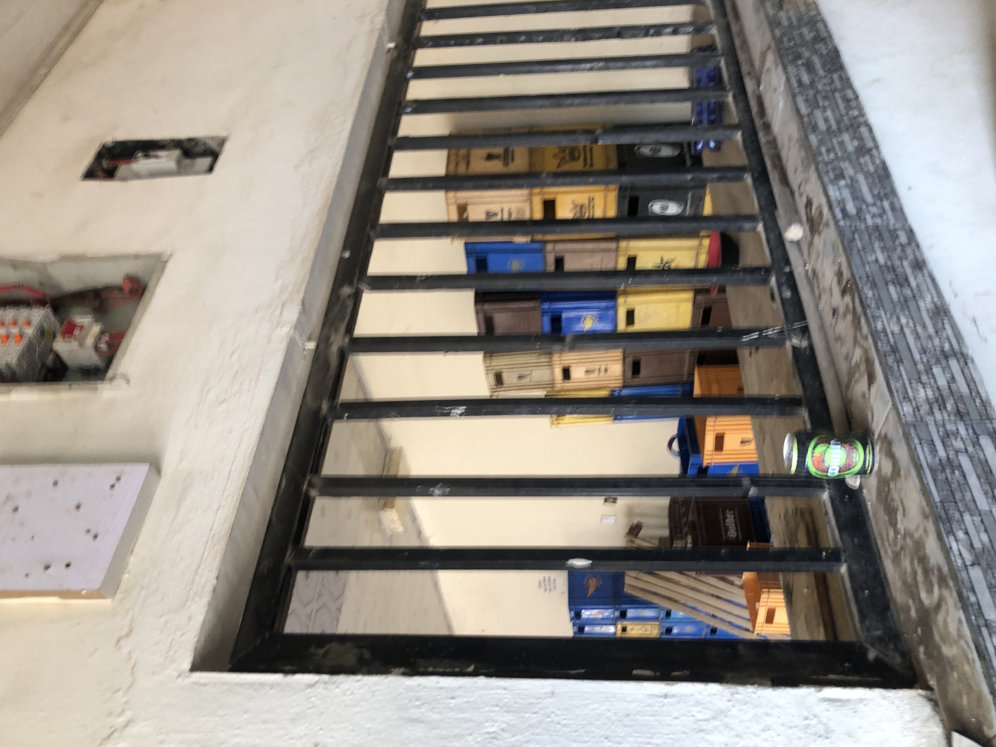 The image presents an angled, downward-looking view into a lower level, likely a storage area or basement, through a black metal grate. The immediate foreground shows a white, roughly textured wall, possibly stucco or painted concrete. This wall features two open rectangular cut-outs: the lower one reveals electrical components including wiring, orange and white circuit breakers, and a fuse box. Below this, a small white board with multiple small dark dots is attached to the wall. The upper cut-out shows less distinct electrical apparatus.
The black metal grate, composed of parallel bars, spans the opening. To the right of the grate, a vertical surface is visible, covered in grey and black patterned tiles. A green and black drink can, with the text "DIET BITTER" faintly visible (possibly Schweppes brand), rests horizontally on one of the grate's bars on the right side.
Through the grate, numerous stacked crates or boxes are visible in the lower area. These crates are of various colors, including yellow, blue, brown, and black, and appear to be plastic beverage crates. Visible text on some yellow crates includes Japanese characters, specifically "麒麟" (Kirin) and "一番搾り" (Ichiban Shibori), alongside "Heineken". Some blue crates also display "Carlsberg".
No people are present, and no specific activity is taking place; the scene depicts a static storage or utility space. The open electrical cut-outs suggest an ongoing project or unfinished work. The brand names on the crates, particularly Kirin, Heineken, and Carlsberg, suggest a commercial setting such as a restaurant, bar, or beverage distribution point. The Japanese text indicates an Asian context. The lighting appears even, suggesting indoor conditions.