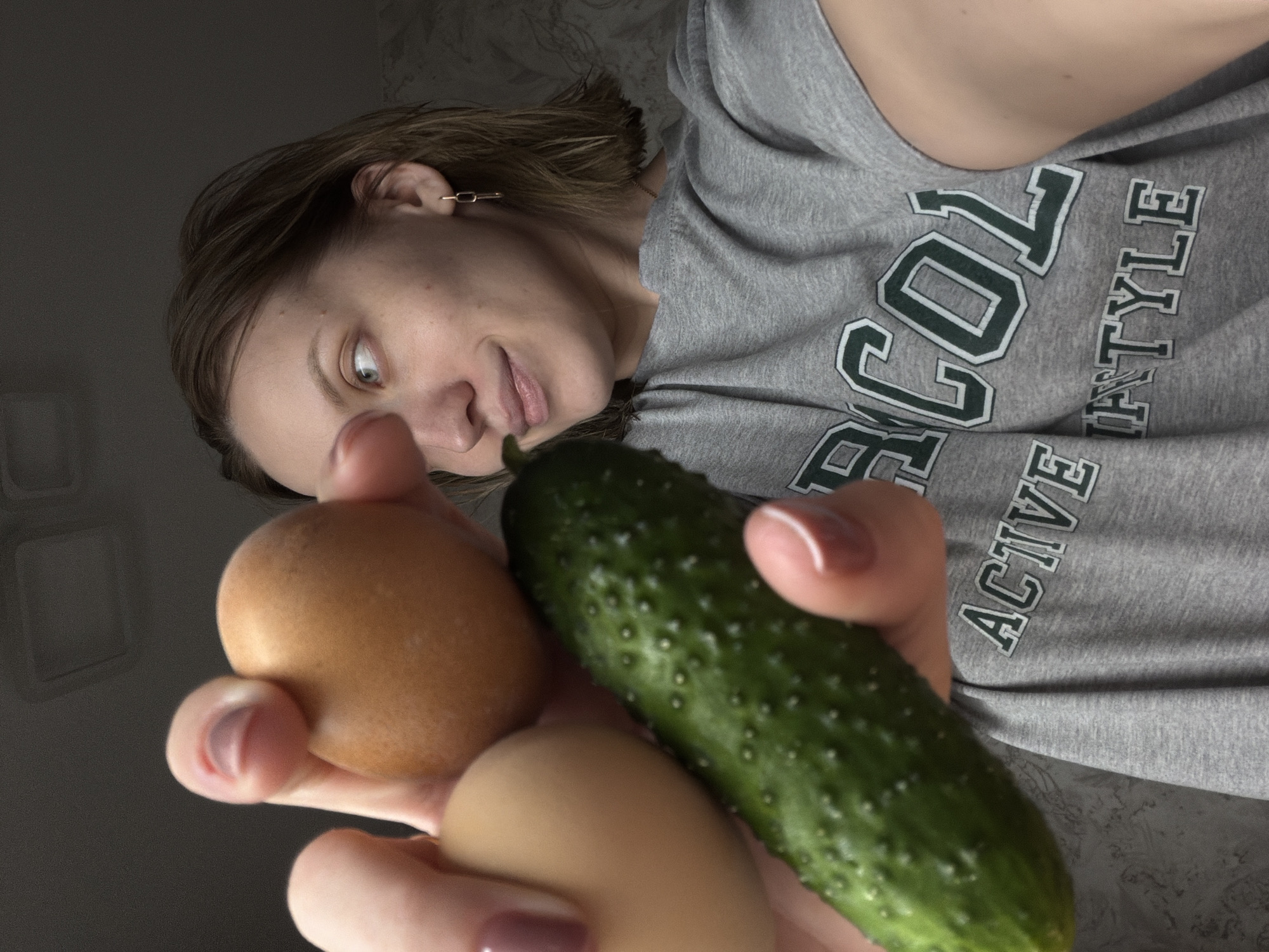 A medium close-up, low-angle shot captures a woman holding a green cucumber and two brown eggs in her hands. Her head and upper torso are visible, rotated counter-clockwise, as she looks down at the items with an expression of curiosity or mild surprise, her eyes wide and mouth slightly agape. She has short, light brown hair and fair skin.
The woman is wearing a grey t-shirt with dark green and white block lettering. Visible parts of the text include "RCOL", "ACTIVE", and "TYLE", suggesting a phrase like "ACTIVE LIFESTYLE" or similar. A small, gold-colored chain earring is visible in her right ear. Her fingernails have a light pink polish.
The background is dark and indistinct, with a faint lighter, possibly patterned, surface visible in the upper left. The lighting is dim, indicating an indoor setting. The image portrays a focused moment of interaction with the cucumber and eggs. This scene takes place in Reshetnikovo, Russia.