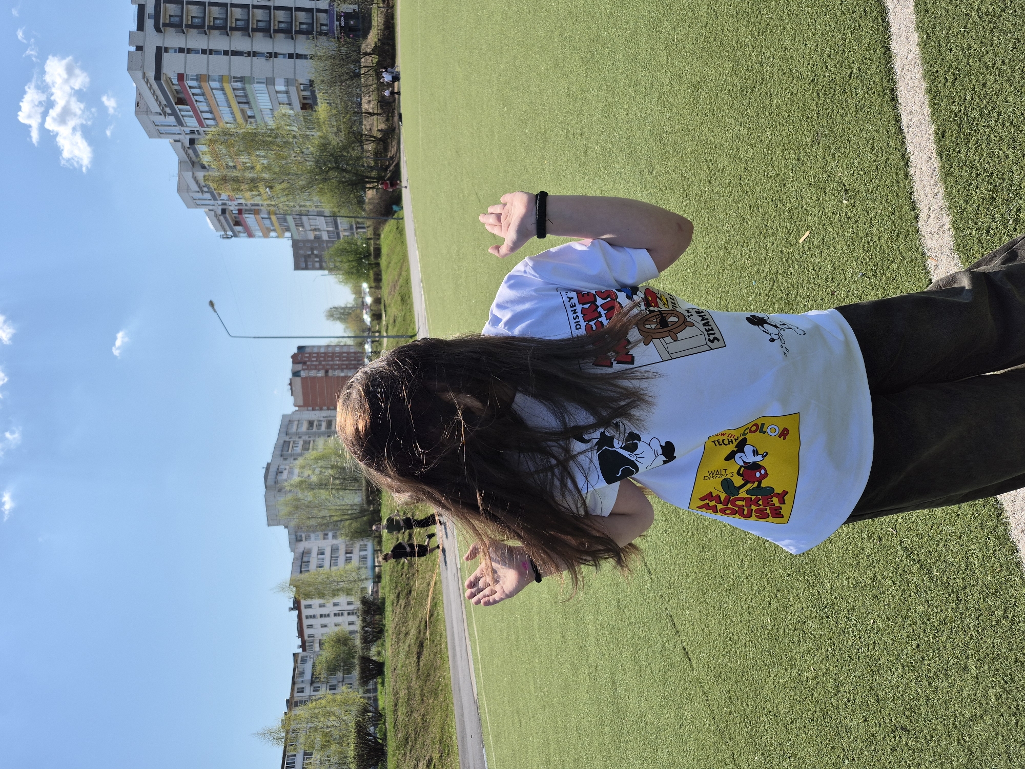 A female stands on an artificial turf field adjacent to a curb and adjacent street with a backdrop of tall residential buildings in Chaikovsky, Russia. She wears a white T-shirt with a Mickey Mouse graphic and dark pants. She is positioned toward the right and appears to be reaching out with both hands. The field, marked with a white line, occupies the top right and bottom right of the frame. A grassy strip with trees and shrubs, separates the field from the buildings. The buildings dominate the upper left of the image, under a partly cloudy blue sky.