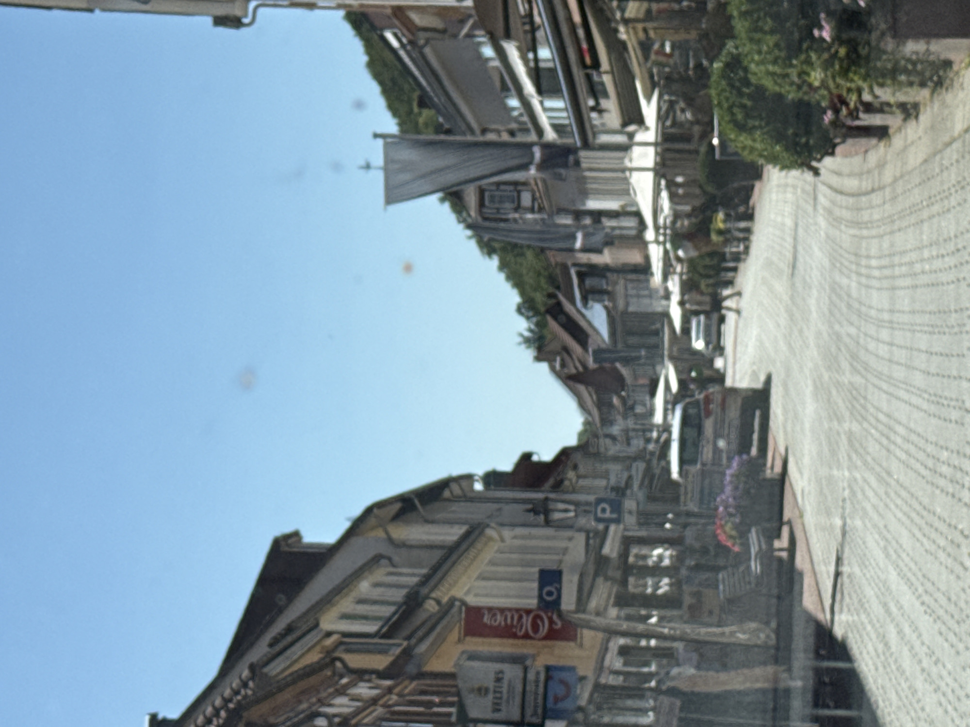 The image depicts a street scene in the historic town of Bad Lauterberg im Harz, Germany, on a sunny day. The cobblestone street curves gently, lined with traditional timber-framed buildings. Shops and cafes with awnings are visible on both sides of the street. In the foreground, a car is parked, and a bench is placed near a planter filled with colorful flowers. People are walking and seated at outdoor cafes, suggesting a lively atmosphere. The background shows a densely wooded hillside, typical of the Harz region. The clear blue sky indicates pleasant weather. The scene captures the charm and typical character of a German town square.