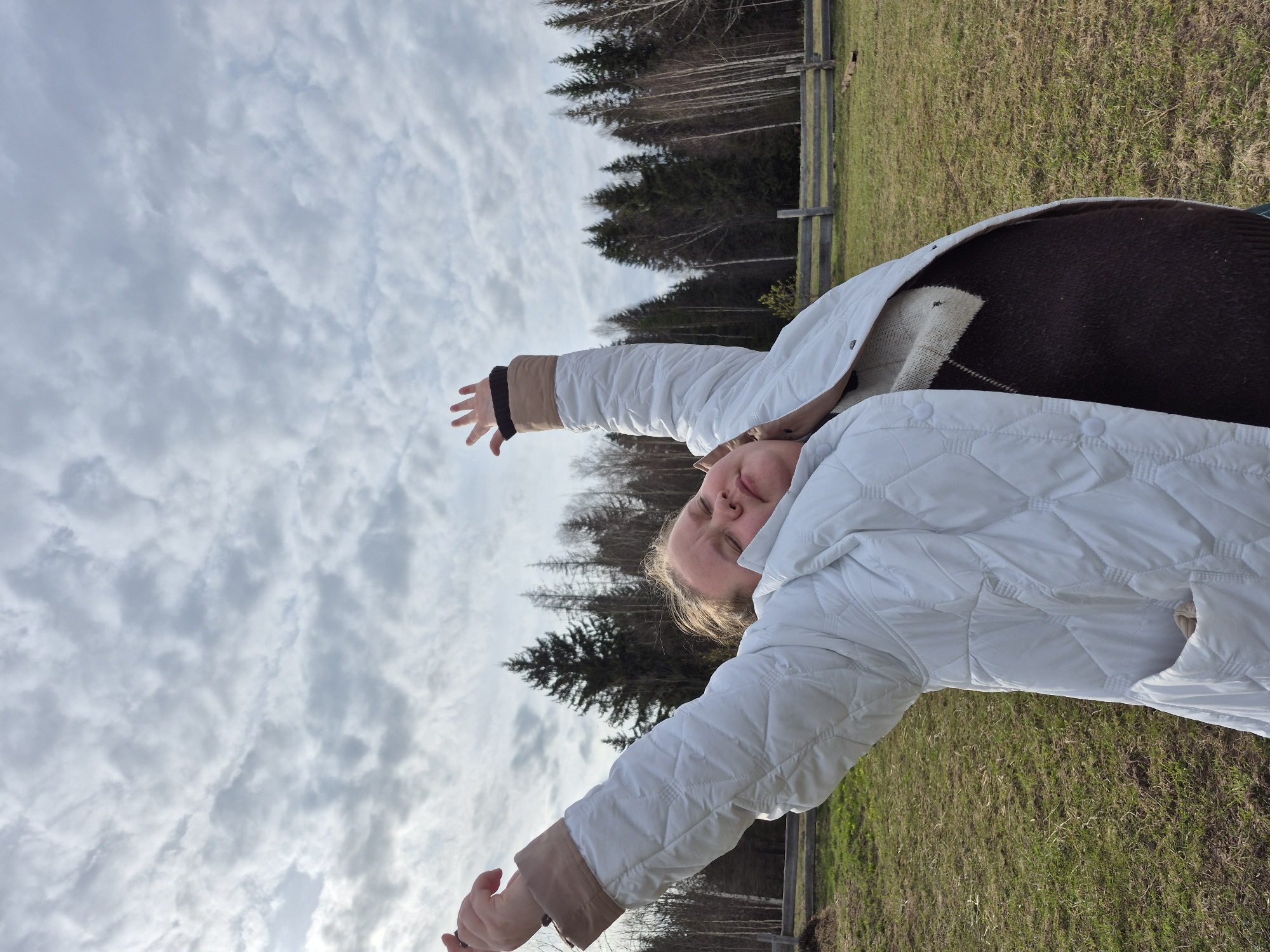 A person is depicted outdoors in a landscape featuring a forest and grassy area, potentially in the vicinity of Дойная, Russia. The individual, wearing a white quilted jacket over a dark sweater, has their arms raised. The background shows a line of trees against a cloudy sky. A wooden fence is also visible. The photograph is taken from a low angle.