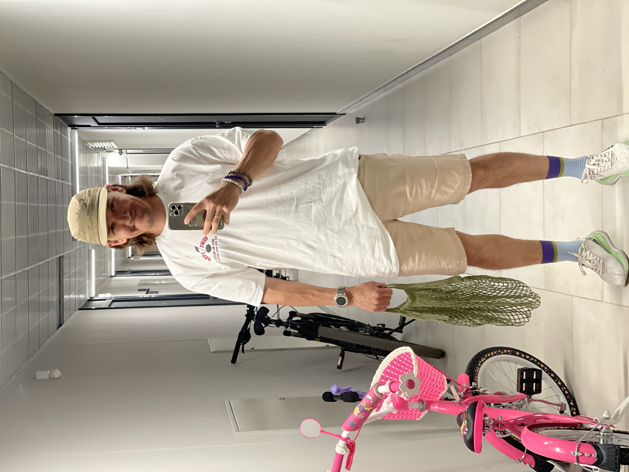 A man wearing a tan baseball cap, white t-shirt, and khaki shorts is taking a selfie in a hallway. He is holding a green mesh bag in one hand and a smartphone in the other. He is wearing a watch and bracelets. One leg is visible, adorned with a light blue sock with a purple stripe near the top and a white athletic shoe with a light green sole. Behind him are two bicycles; one is adult-sized and dark colored, and the other is a pink child's bike with a white basket. The hallway has white tiled walls and floor, and a dark horizontal stripe near the ceiling. The location is Izhevsk, Russia.
