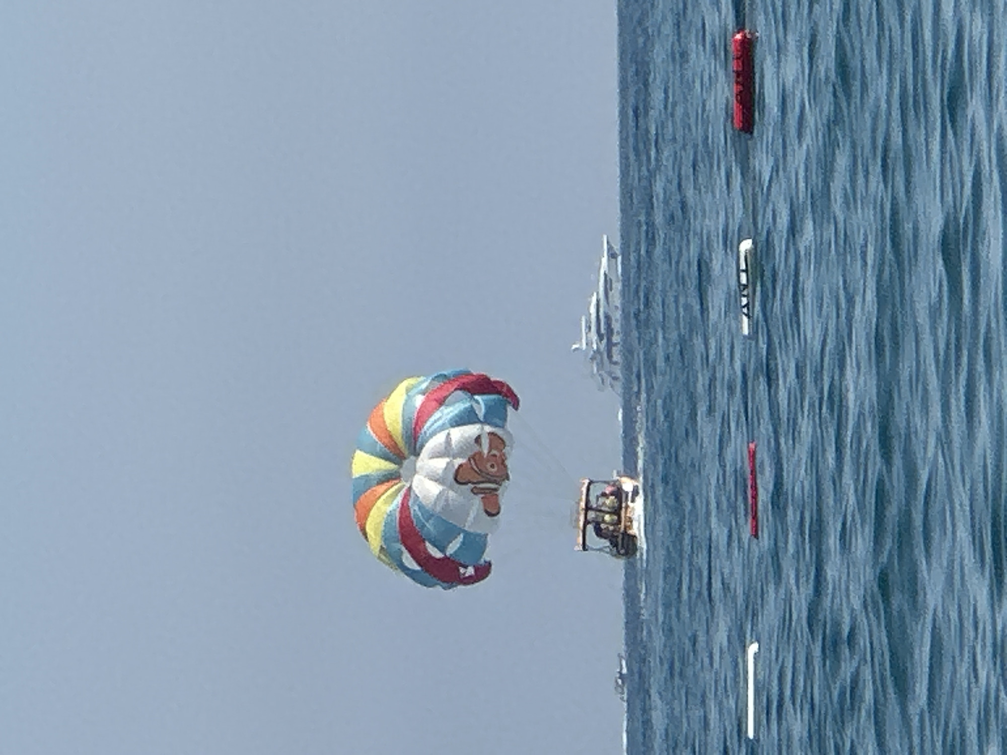In the city of Boreti, Montenegro, a brightly colored parasail with an image of the cartoon character Nemo is visible in the sky above a boat on the azure waters of the sea. The boat appears to be towing the parasail, carrying people who are enjoying the activity. In the background, a large white yacht is anchored, and other smaller boats are scattered across the sea. The horizon is clear, with a pale blue sky overhead. Several buoys, some red and white, are floating on the surface of the water in the foreground.
