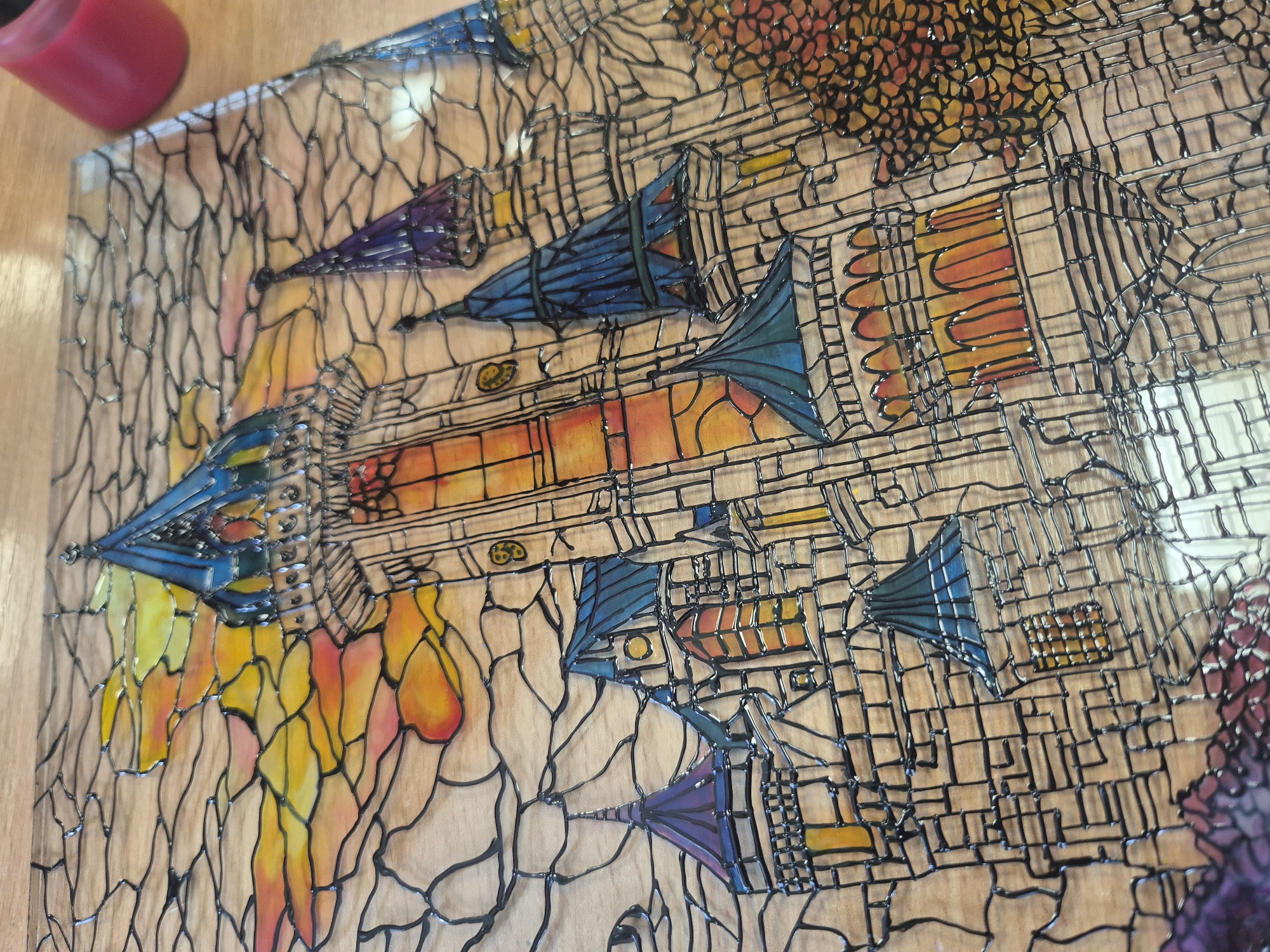 The image shows a glass artwork depicting a fantasy city. The artwork has a grid-like, black outline that separates different sections of the city. The colors used are vibrant, including blue, purple, orange, red, and yellow. The city appears to be a mix of buildings and trees. The artwork is sitting on a light brown wooden surface. A red candle is located in the upper left corner. The location, Pavlikeni, Bulgaria, is not represented in the image.