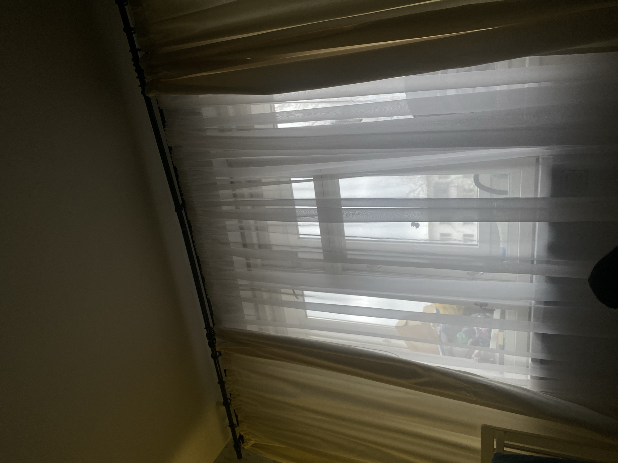 A daytime interior scene in Chaikovsky, Russia, features a window with layered curtains, a sheer white layer and heavier gold/beige layer. The window provides a view of buildings and trees outside. There is a dark object on the right side, and the edge of some furniture appears at the bottom. The left side of the frame is a wall, with dark curtain rods next to it.