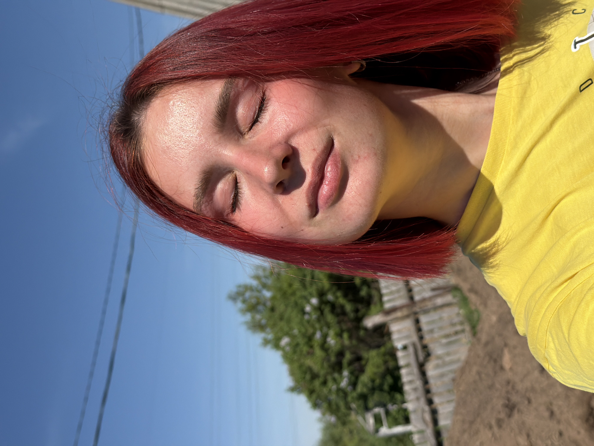 A young woman with red hair and closed eyes is shown in an outdoor setting. She is wearing a yellow shirt. The background includes a bright blue sky with power lines, trees, and a wooden structure or fence. The image appears to be taken in daylight. There is no confirmed presence within the city of Chaikovsky, Russia.