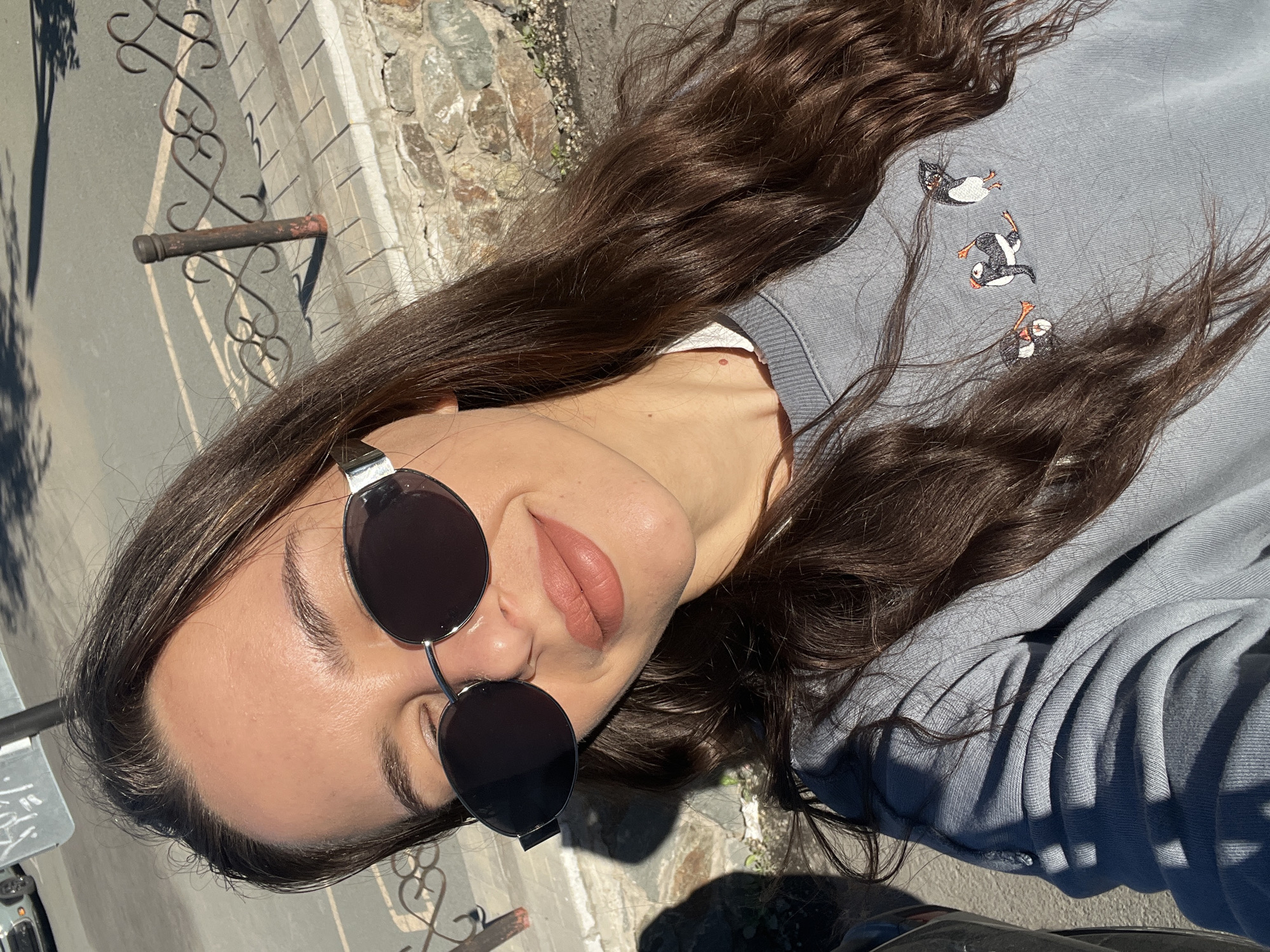 A woman is pictured wearing sunglasses and a light blue sweatshirt with puffin designs. She has long, dark hair. The background includes a cobblestone wall, a decorative metal fence, and a partially visible street. The location appears to be in Vladivostok, Russia, suggested by the architectural style and layout.