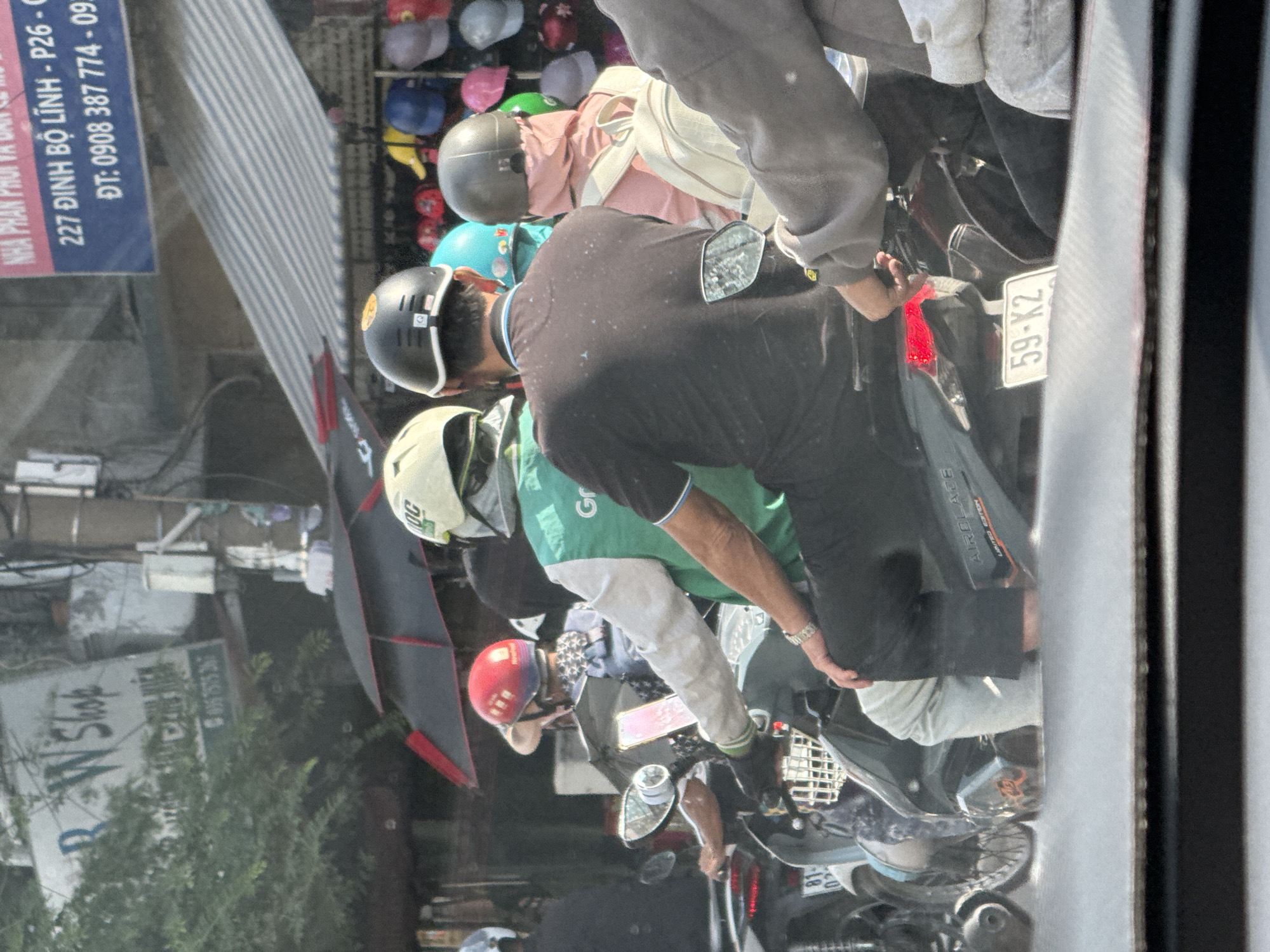 The image captures a street scene in Thủ Đức City, Vietnam, likely during daylight. In the foreground, a person wearing a gray jacket is seated on a scooter, with their right arm extended slightly. Behind them, another individual in a black shirt and light-colored pants is also on a scooter, with a mobile phone mounted on the handlebars. To the left, a person in a red helmet and a light-colored hat is on a scooter, holding a smartphone. Another scooter rider, wearing a green vest and a white helmet, is partially visible. Multiple other individuals on scooters are present, some with helmets, suggesting a busy traffic environment.
In the background, shops are visible, with awnings and displays of merchandise, including hats of various colors. A sign with Vietnamese text is mounted on a building. An umbrella is deployed, providing shade. The overall environment appears to be an urban street with commercial activity. The license plate on the scooter in the foreground reads "59-K2".