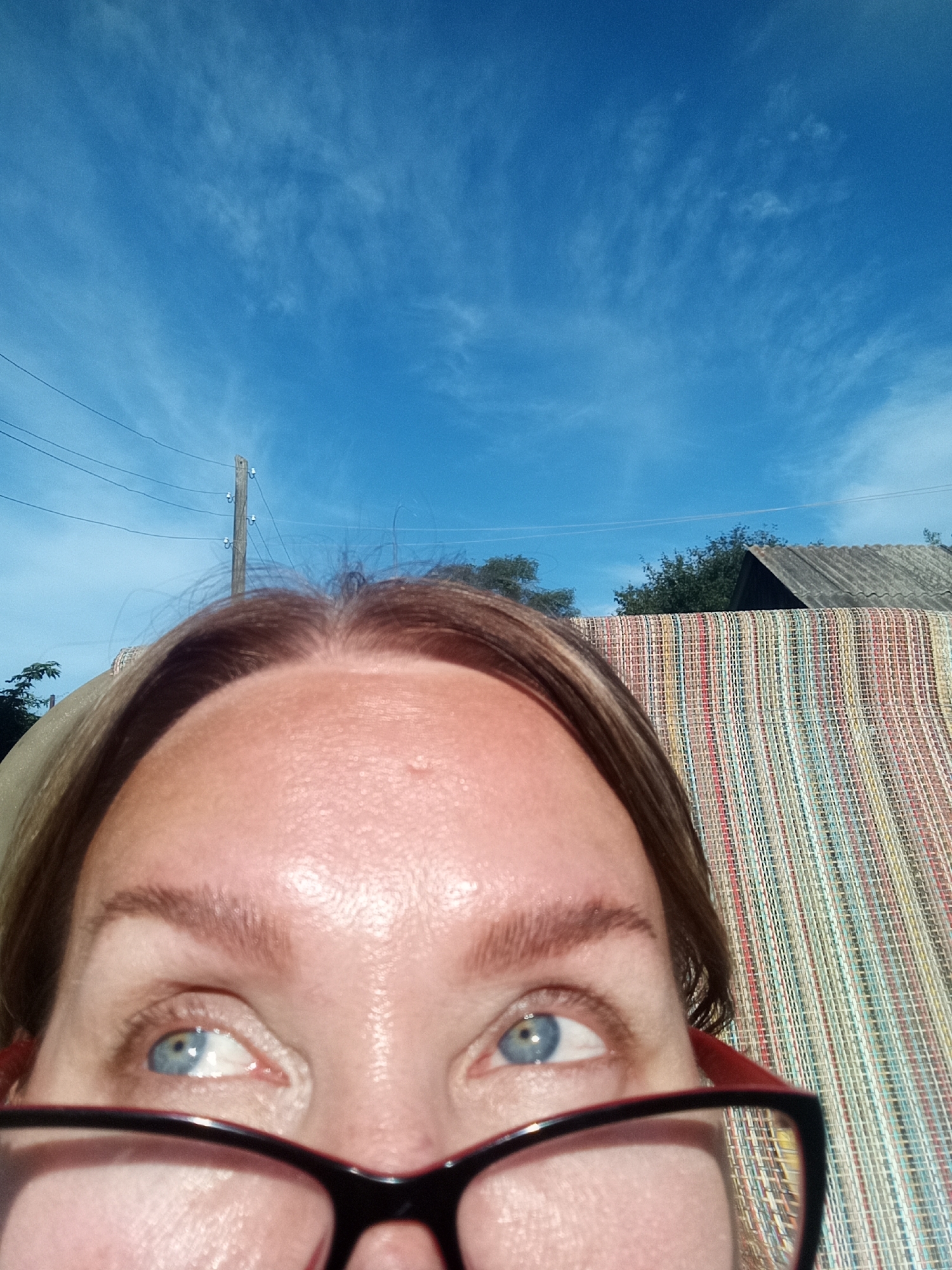 An extreme close-up shot shows a person's forehead and eyes, wearing red-rimmed glasses. The person's eyes are blue and looking upwards. The background features a bright blue sky with wispy clouds, a utility pole with wires, trees, and a rustic building with a corrugated roof. The scene appears to be outdoors, possibly in a rural or suburban area of Novopokrovka, Russia. A textured, colorful mat is visible behind the person's head.