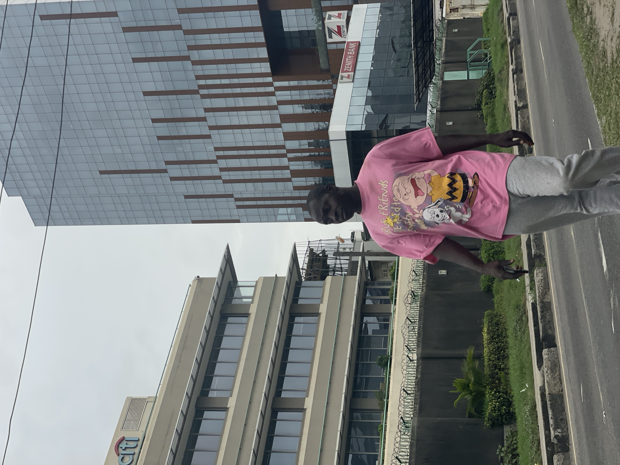 A pedestrian stands on a roadside in Itirin, Nigeria, adjacent to two prominent buildings. One building is a modern glass-clad structure, identified as Zenith Bank. The other is a taller office building with a logo that reads "Citi." The scene features typical urban elements such as a road, concrete curbs, landscaping with shrubs and trees, and a concrete wall topped with coiled barbed wire. The sky is overcast. The individual wears a pink t-shirt with cartoon characters and grey pants.