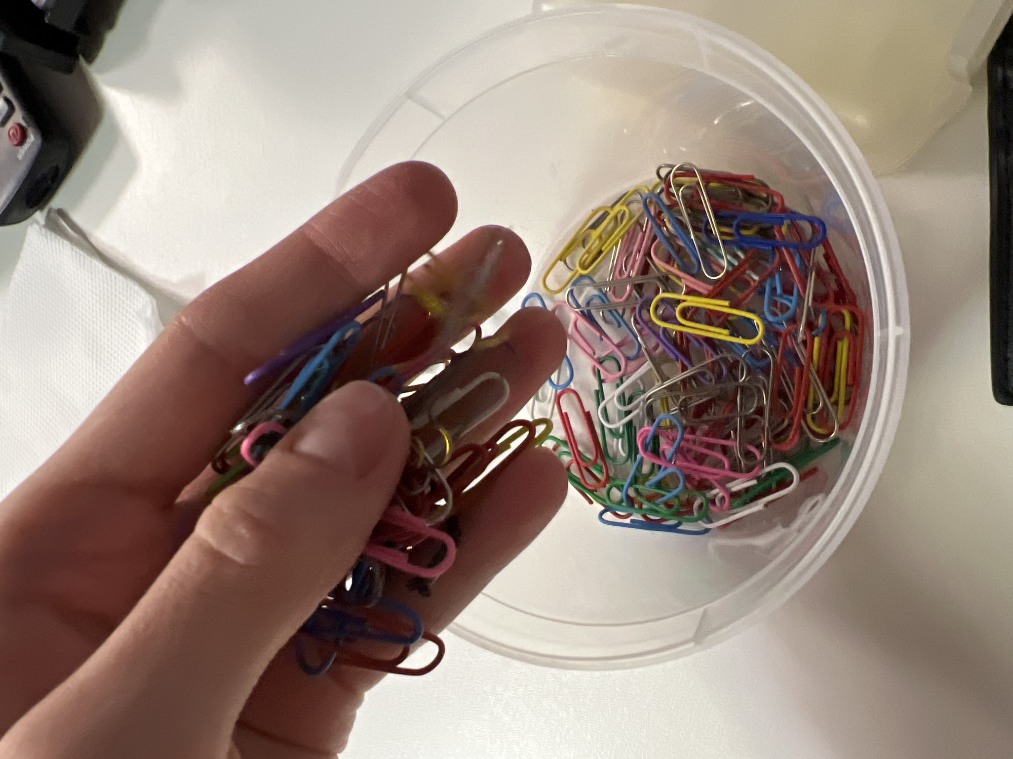 A close-up, high-angle shot captures a human hand interacting with colorful paper clips on a light-colored surface. A fair-skinned hand, likely an adult's, holds a cluster of paper clips in the foreground, with individual clips in shades of blue, purple, yellow, pink, red, green, and silver/white visible between the fingers.
To the right, a clear, round plastic container is filled with a larger assortment of similarly multi-colored paper clips, jumbled together. The hand appears to be in the process of either taking paper clips from or placing them into this container.
The scene is set indoors, likely on a desk or workstation, as indicated by the presence of office supplies and a predominantly white, smooth surface. In the upper left corner, a portion of a black electronic device with red buttons, possibly a camera or remote, rests on a white tissue or napkin. The lighting is bright and even, suggesting a well-lit interior during the day or under artificial light. There is no visible text in the image. The activity suggests organizing or preparing to use office supplies in an everyday setting in Khimki, Russia.