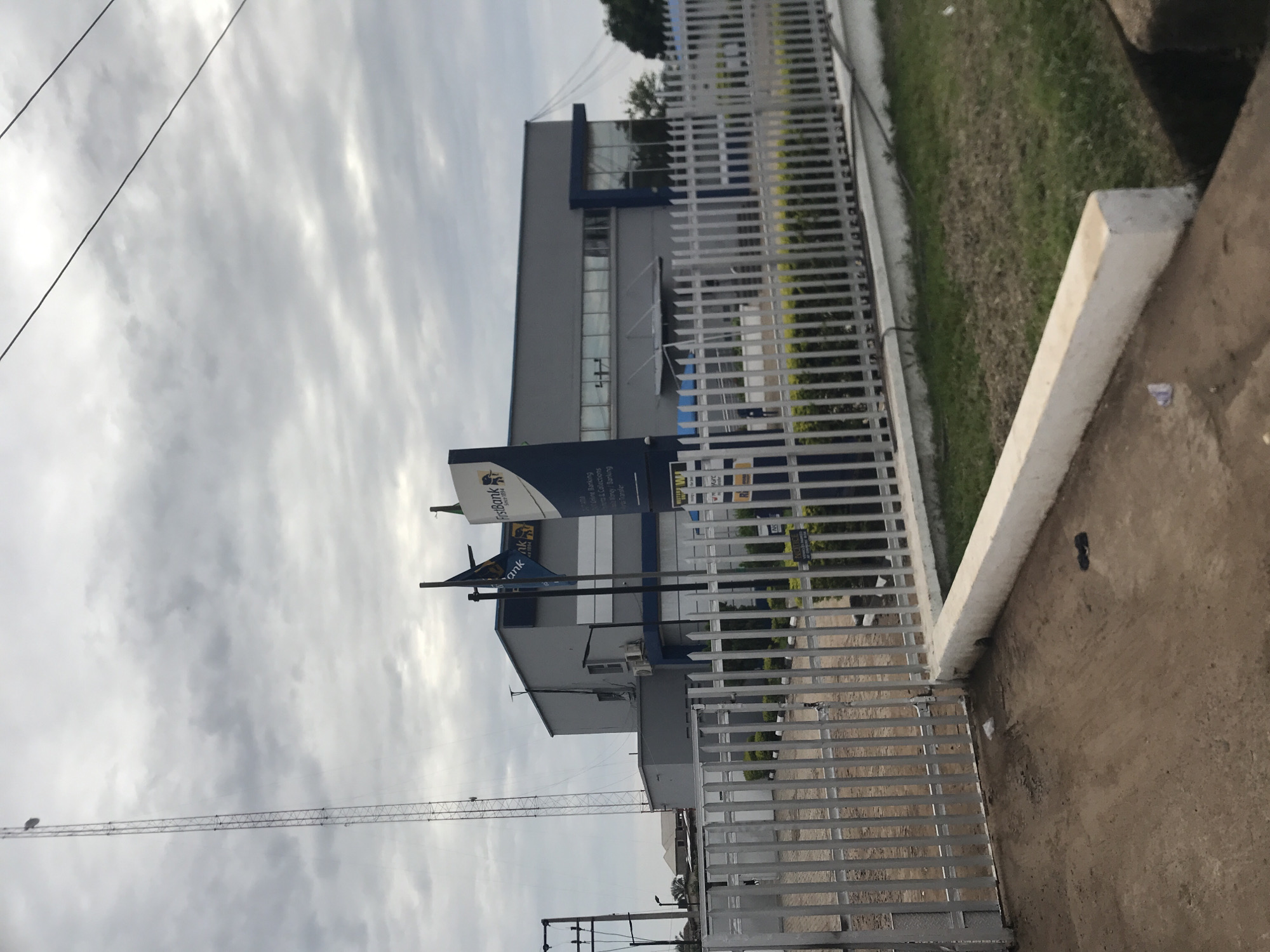 This image captures an exterior view of a FirstBank branch located in Rafin Par, Nigeria, under an overcast sky. The primary subject is a two-story building constructed with grey facades and prominent blue architectural accents, featuring large reflective windows on the upper level. A large, freestanding sign with the FirstBank logo (an elephant emblem) and text detailing banking services, including "Online Banking," "Funds & Collections," "Money Transfer," and "Western Union," is positioned to the right of the building's entrance. A flag bearing the FirstBank logo is flown on a pole adjacent to the sign.
To the left of the bank building, a very tall and slender lattice communication tower extends high into the grey, cloudy sky. Overhead utility lines are visible crossing the upper part of the frame and on the left side connected to a utility pole.
In the foreground, a tall, silver-colored metal fence composed of vertical bars and horizontal supports runs across the entire width of the scene, with a gate section present on the left. A low, white concrete barrier runs along the base of the fence on the viewer's side, transitioning into a dirt and concrete ground surface on the left. Beyond the fence, the bank property features a manicured lawn with interspersed green shrubs. Litter, including a small white plastic item and a dark object, is visible on the concrete ground in the immediate foreground. No individuals are present in the scene. The sky is entirely covered with dense grey clouds.
