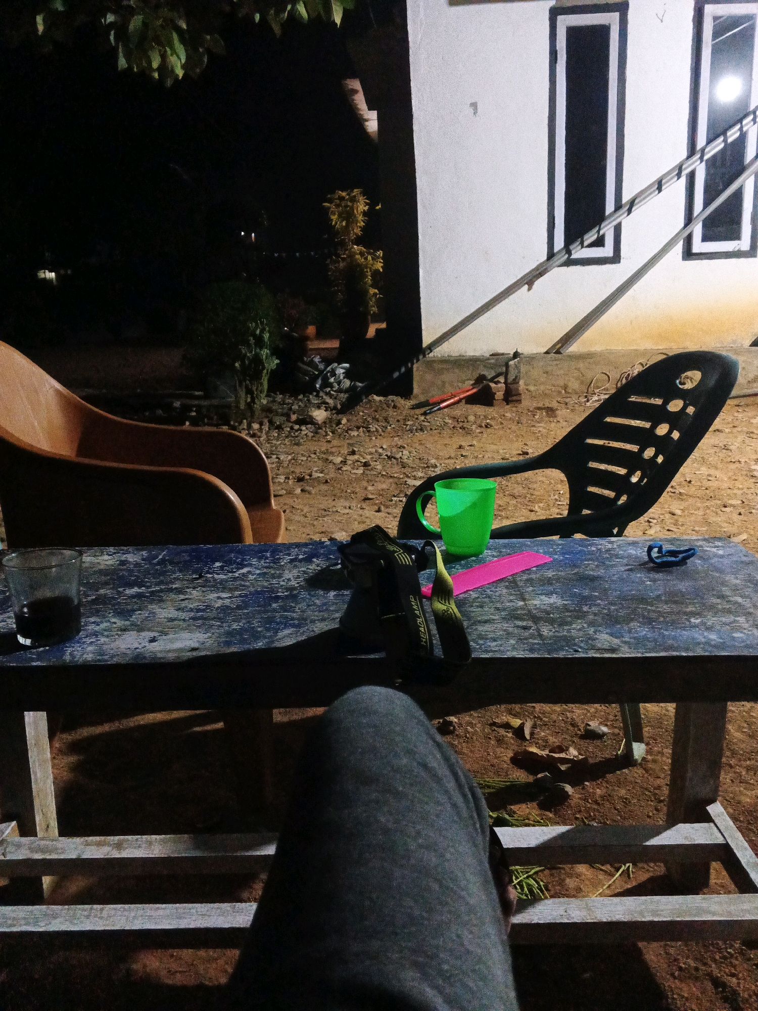 A low-angle, eye-level shot captures a dimly lit outdoor scene in what appears to be a rural area, possibly Brangan Mulya, Indonesia. The foreground is dominated by the legs of an unseen individual, clad in grey sweatpants, seated on a rustic wooden bench. In front of the bench is a weathered, dark blue wooden table, upon which rests a clear glass containing a dark liquid, likely coffee. To the right of the glass, a green plastic mug sits near a pair of dark sunglasses. Further right on the table, a bright pink object, possibly a notebook or a folder, is partially visible, alongside a blue cord.
Behind the table, a black plastic chair with an intricate, perforated design is positioned. The ground surrounding the table and chairs is dirt and gravel, with scattered leaves and small stones. In the background, a white building with two tall, dark-framed windows is visible, with a metal railing system ascending beside it. The overall ambiance is one of quiet nighttime relaxation in an outdoor setting.