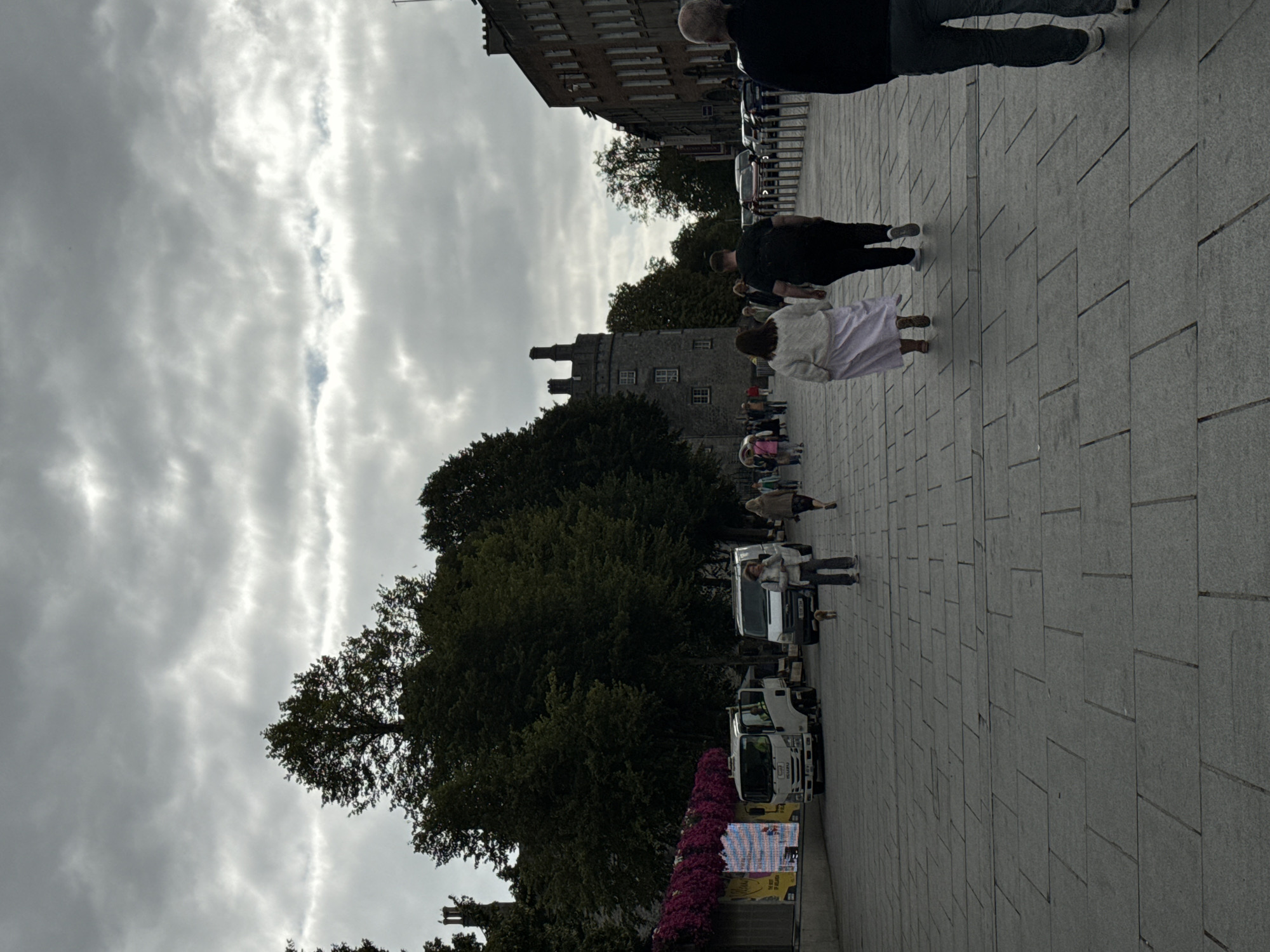 The image depicts a street scene in Kilkenny, Ireland, likely near Kilkenny Castle, with a cobblestone pavement and overcast sky. In the foreground, a person walks away from the camera. Further down the street, a couple walks towards the camera, the woman wearing a light-colored dress and the man in dark clothing. Several other people are visible walking and standing. To the left, a white truck and a white van are parked. Behind them, trees and a building with pink flowers are seen. In the background, a stone tower, part of the castle, stands tall. The overall atmosphere suggests a slightly cool, cloudy day in a historic urban setting.